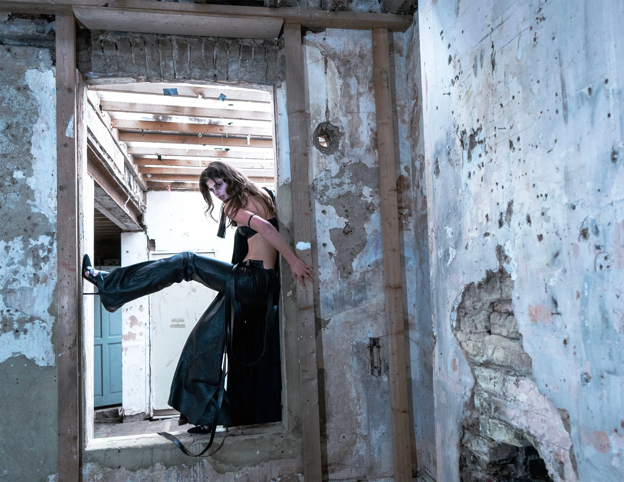 A woman in black leather pants and an open jacket revealing her midriff, sitting on a wooden beam in an unfinished room with exposed brick, wooden studs, and peeling paint.
