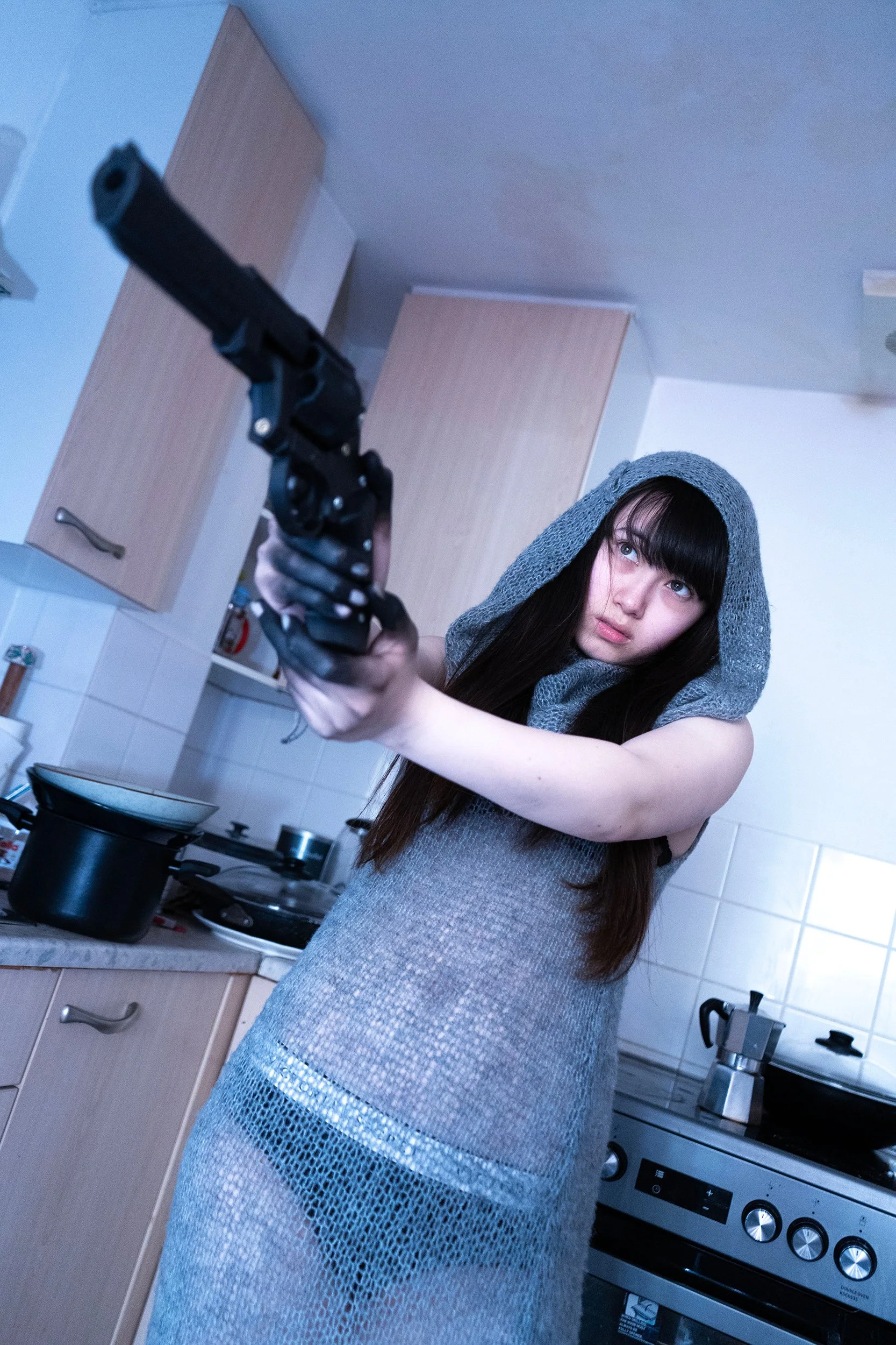 Young woman with long dark hair wearing a gray hoodie and a shiny, metallic skirt, pointing a handgun toward the camera in a kitchen with a stove, pots, and wooden cabinets.