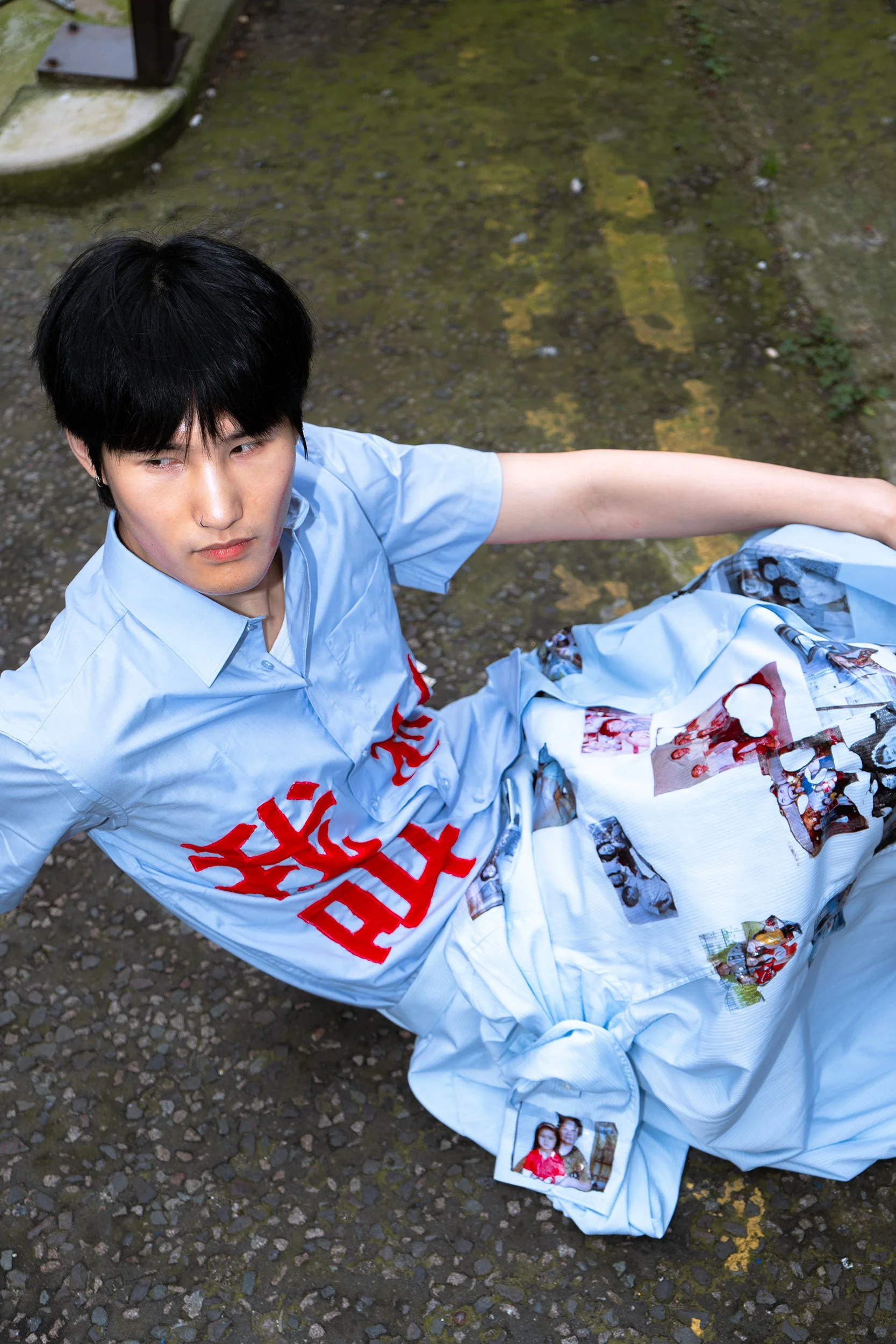 A young man with black hair, wearing a light blue shirt, sitting on a rocky ground with moss. He is looking to his left. There are photos and texts on his white fabric clothing.
