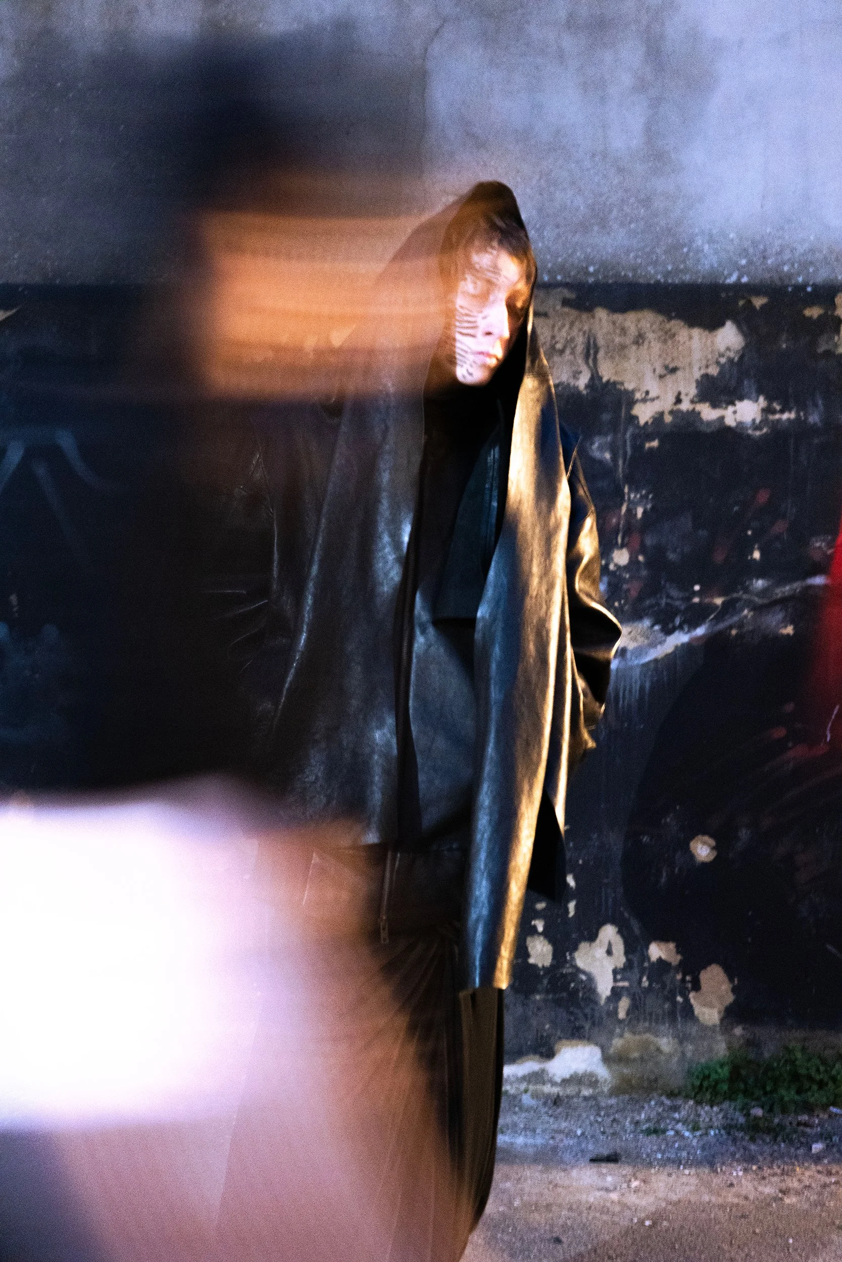 Person wearing a leather jacket and face paint, looking to the side, standing in front of a weathered wall.