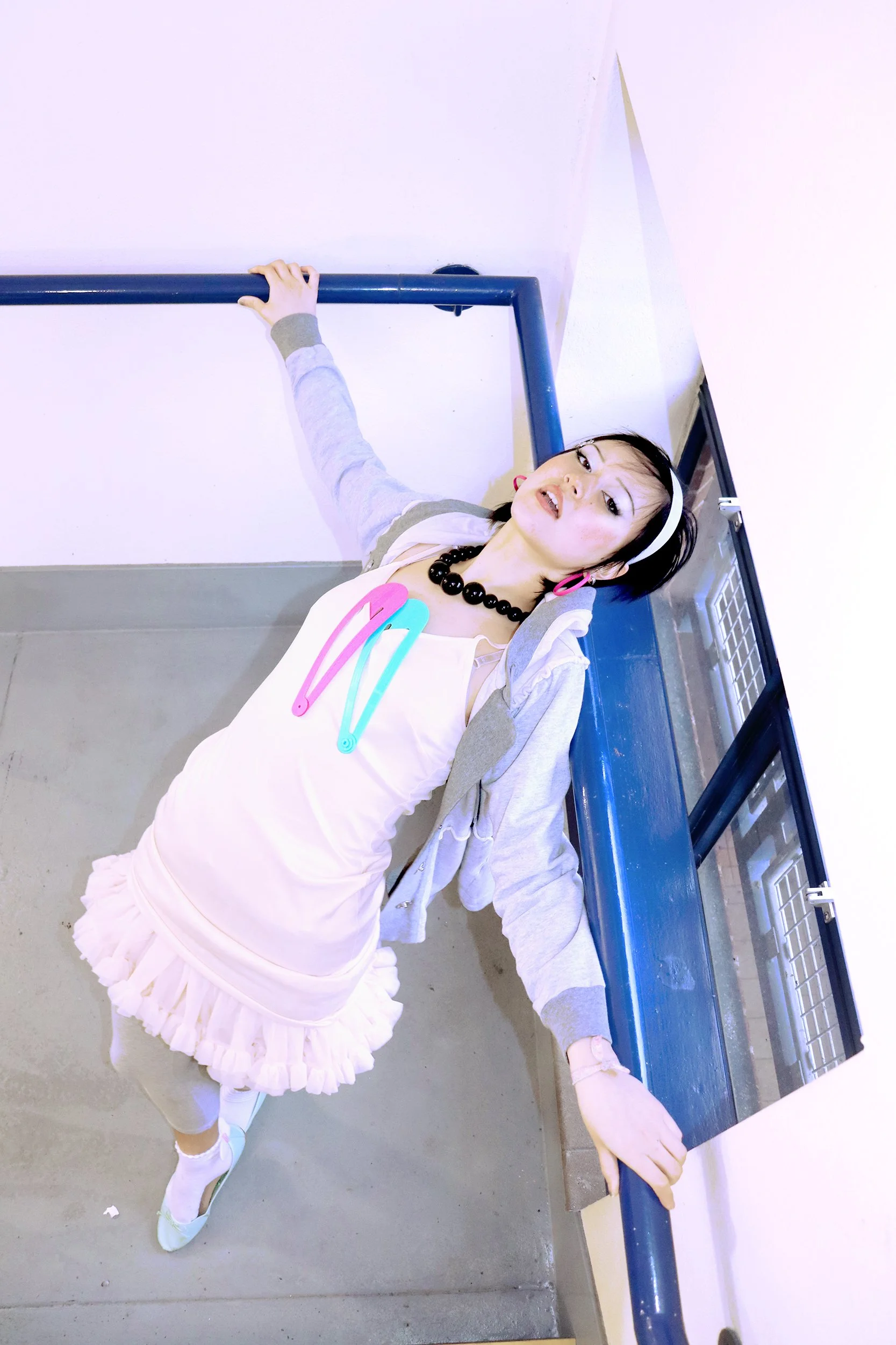 A young woman lying on the floor, reaching out and holding onto a blue railing, wearing a layered pastel pink and white dress with ruffles, a gray zip-up hoodie, white socks, and pastel pink shoes. She has a black beaded necklace, a white headband, and pink earrings.