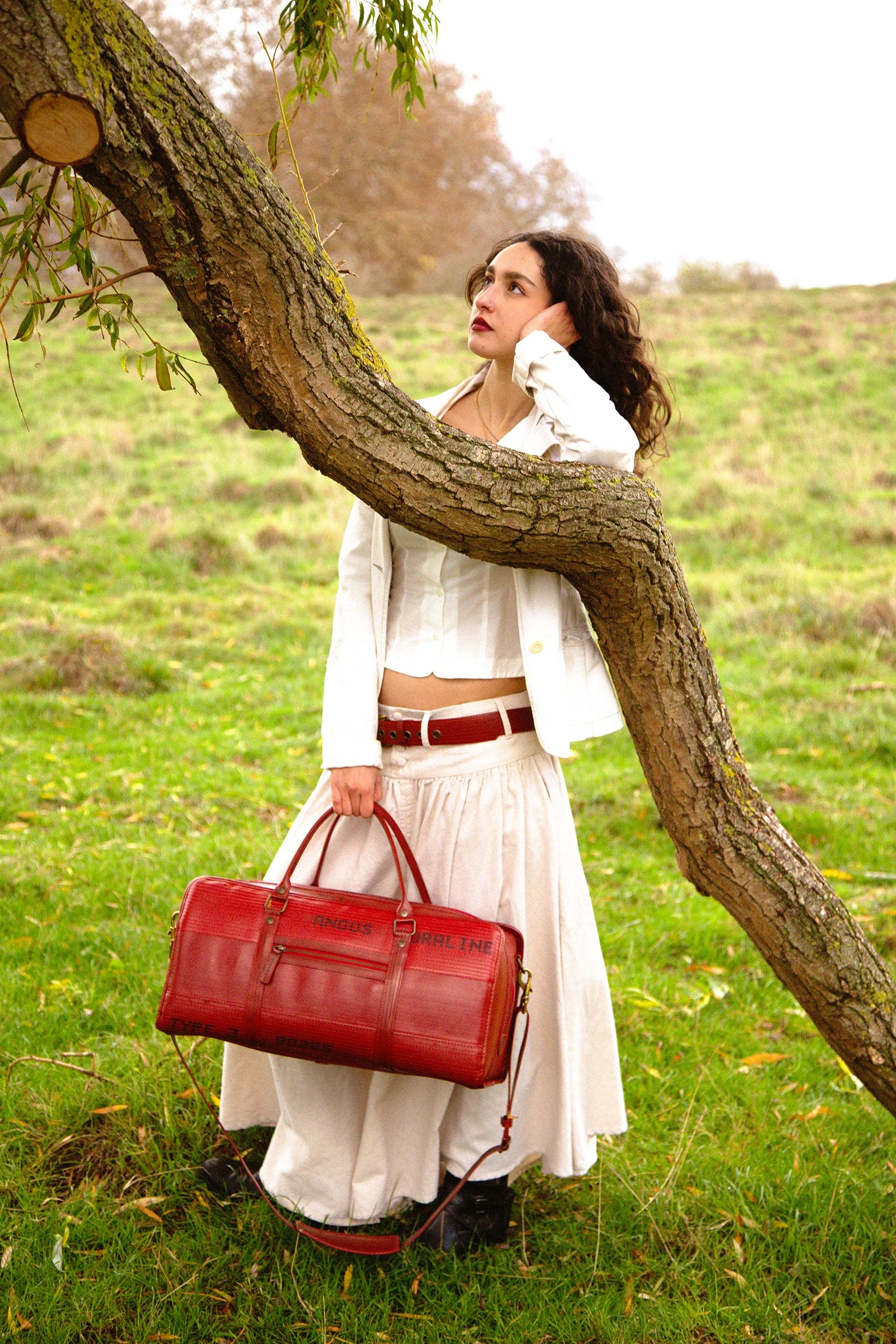 A woman with curly dark hair wearing a white jacket and a white skirt holding a red duffel bag standing outdoors next to a large bent tree with green grass and some fallen leaves, overcast sky in the background.