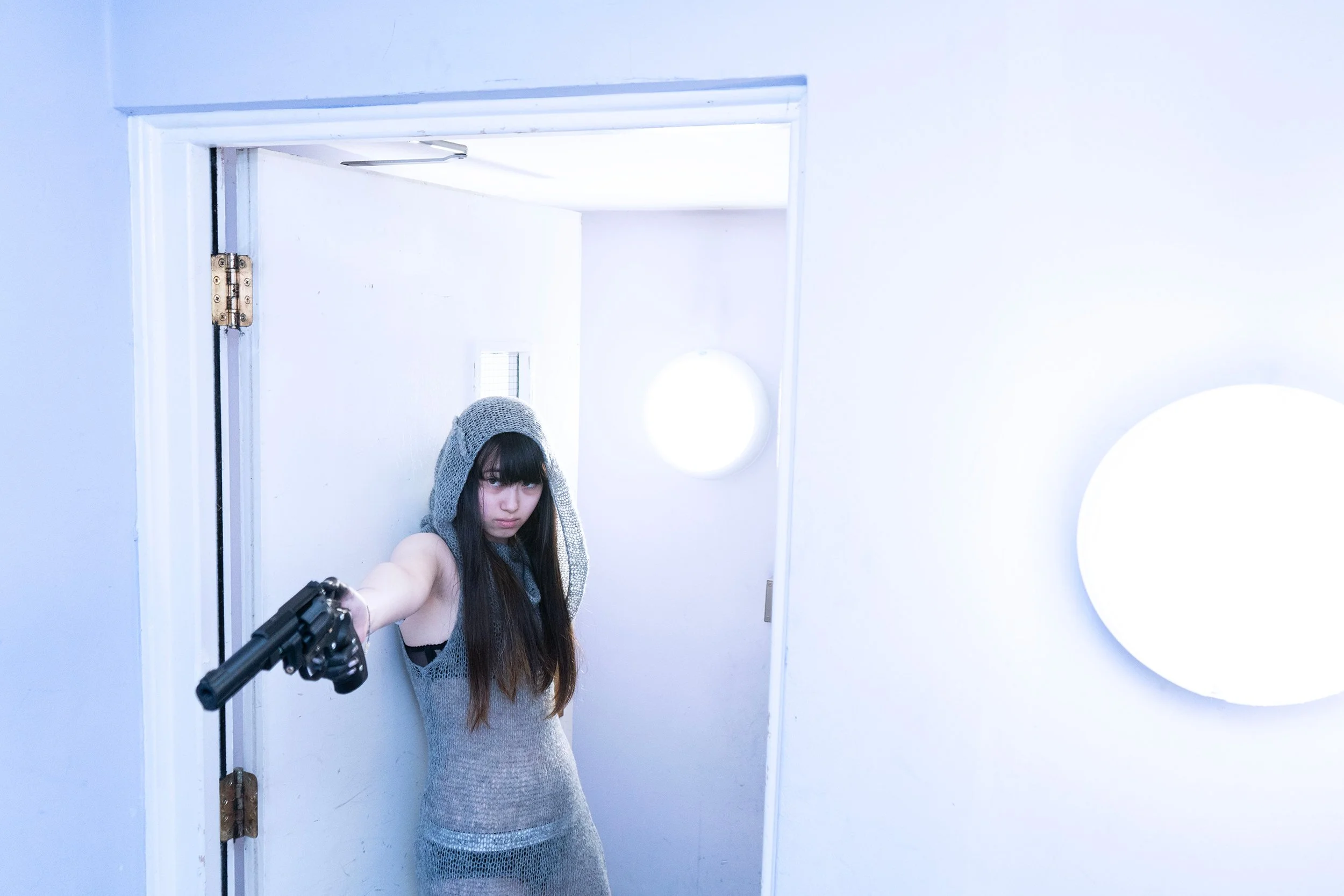 A woman with long dark hair and a gray hoodie is pointing a gun at the camera while standing in a small room with white walls. She appears to be taking a selfie in a mirror, and the room has minimal decor with a small illuminated round wall light and a circular mirror.