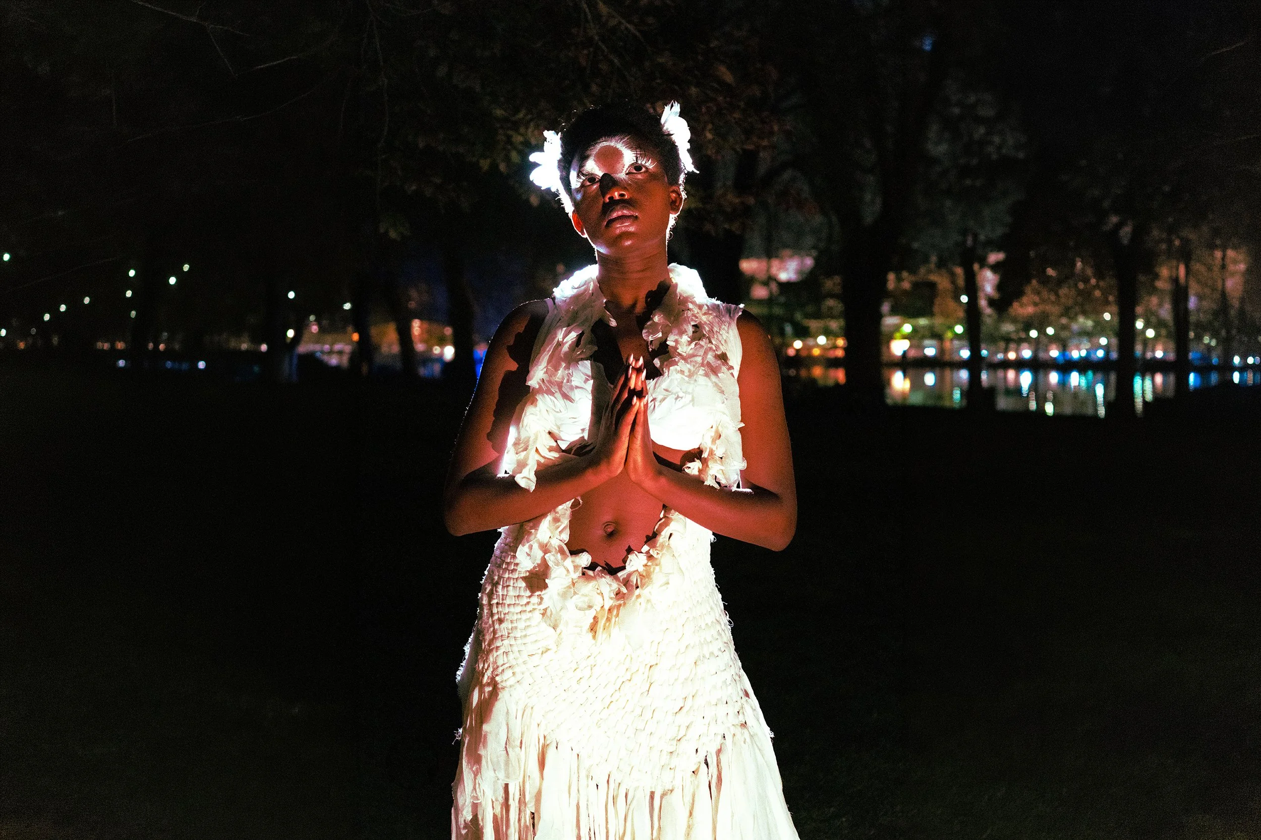 A person stands outdoors at night, wearing a white textured dress with floral details, and a matching headpiece with feathers. They are praying or meditating with their hands pressed together in front of their chest, and their eyes are closed. The background features a dark park with trees, and colorful city lights reflecting on a body of water.