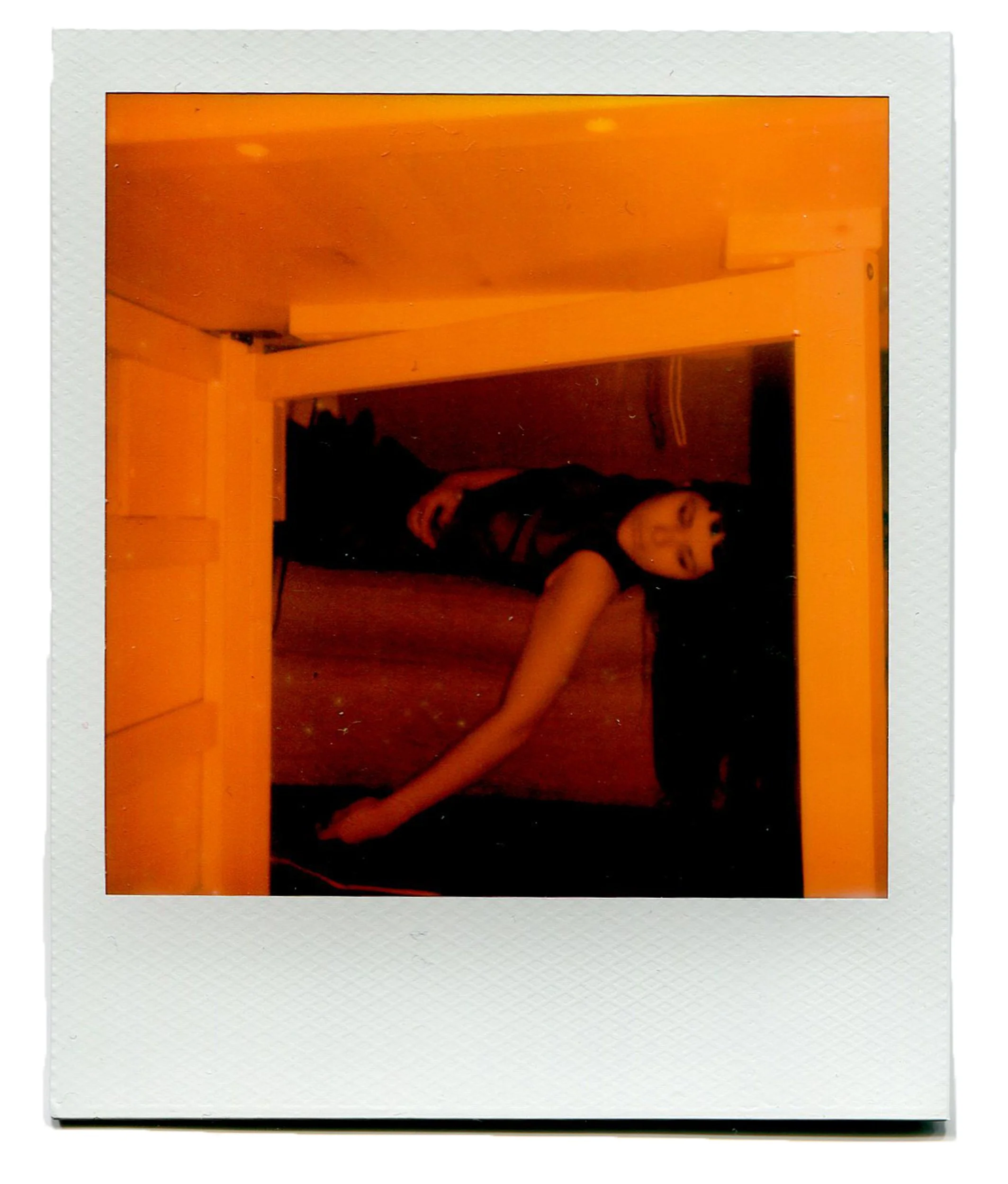 Polaroid-style photo of a woman with dark hair lying on a floor in a room with yellow-orange lighting, with her eyes closed and head resting on her arm.