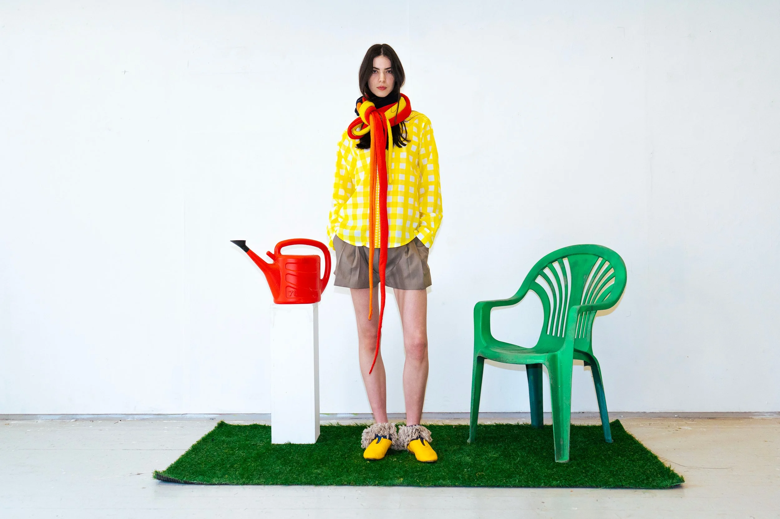 A young woman stands on a small patch of artificial grass, wearing a yellow checkered shirt, beige shorts, and yellow slippers with furry ankle cuffs. She has long dark hair and a serious expression. There is a green plastic chair to her right, a red watering can on a white pedestal to her left, and a black chair partially visible on the left side of the image. She has a colorful scarf wrapped around her neck and hanging down in front.