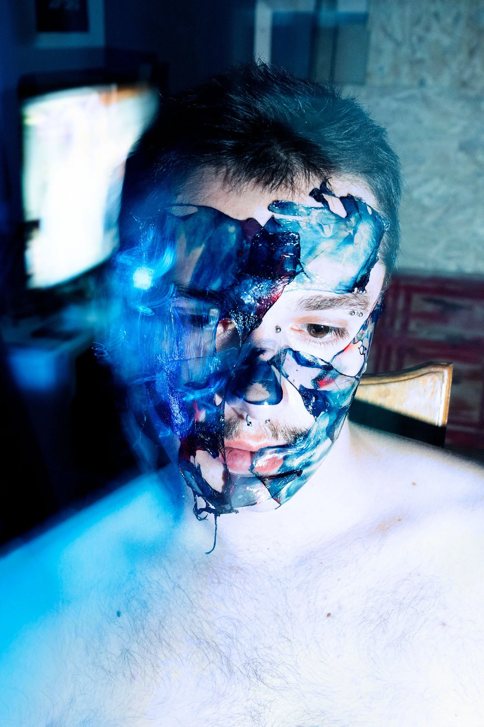 A person with face paint resembling a skeleton or monster, wearing a plastic mask, with dark hair, sitting at a table. The background shows a television, and the image has a bluish hue.