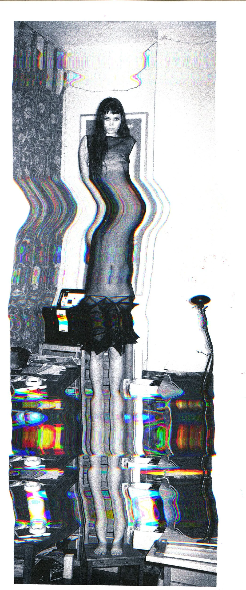 A black and white photo of a woman standing on a table, with a distorted, glitch effect. She is wearing a dress and has long dark hair. The room appears to be an indoor space with a wall, a corner, a small desk with various objects, and a standing lamp.