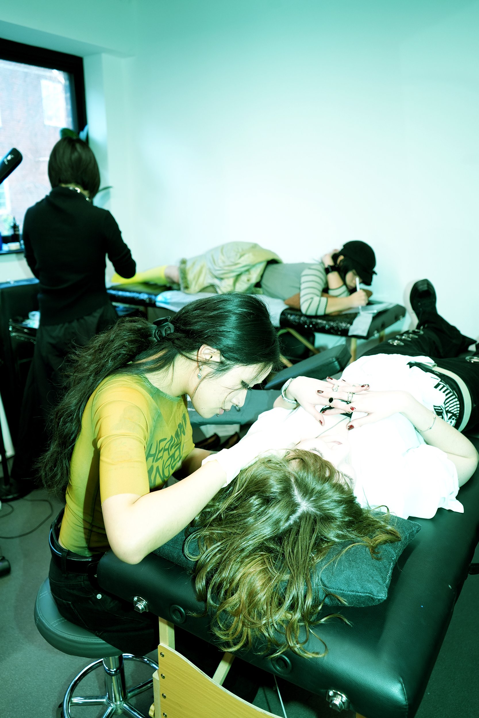 Tattoo artist in a yellow-green top working on a client lying on a tattoo table in a studio, with another person in the background.
