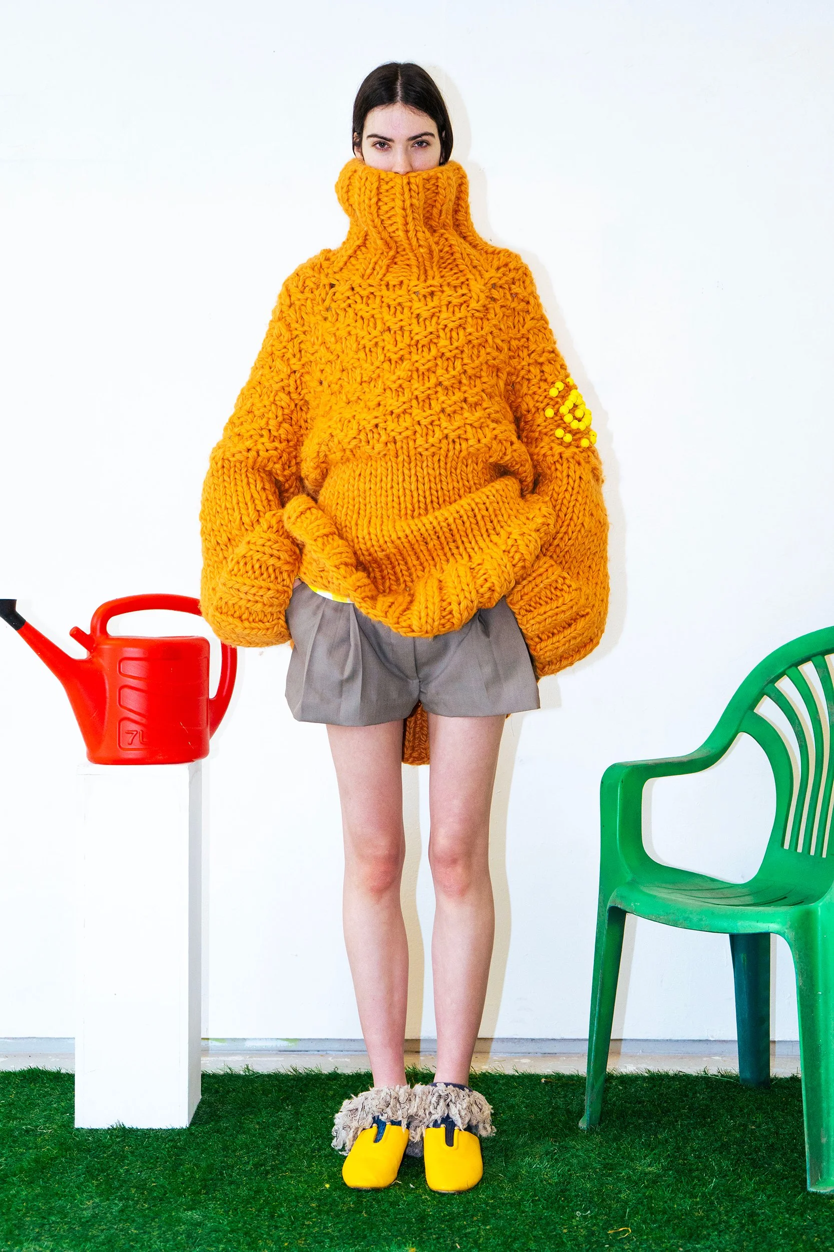 A woman standing indoors against a white wall, wearing a large orange knitted sweater with turtleneck pulled up over her face, gray shorts, furry slippers, and yellow rubber boots. Green and red garden chairs and a red watering can are visible around her.