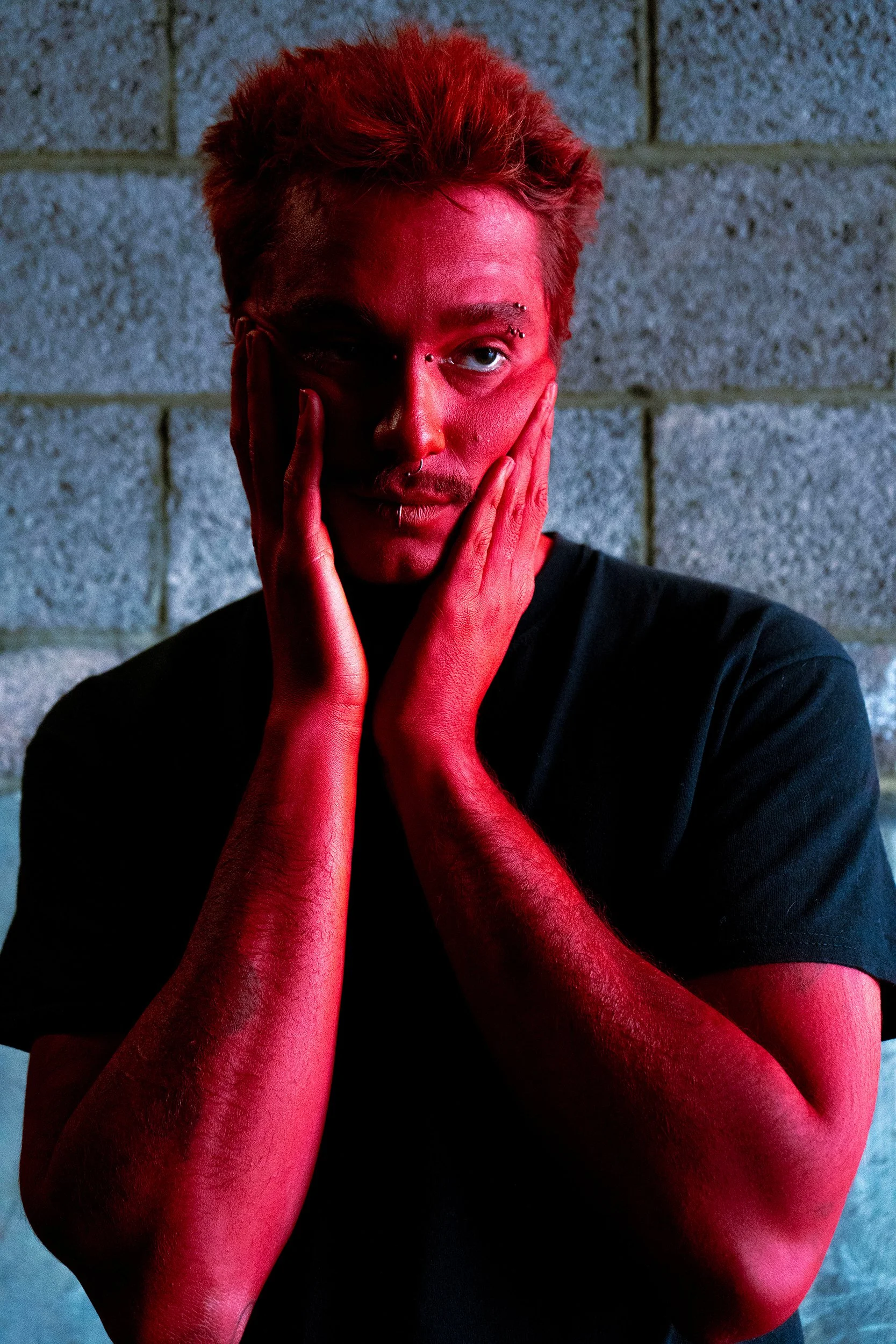 A person with ginger hair and multiple facial piercings, wearing a black shirt, holding their face with hands painted red against a gray brick wall background.