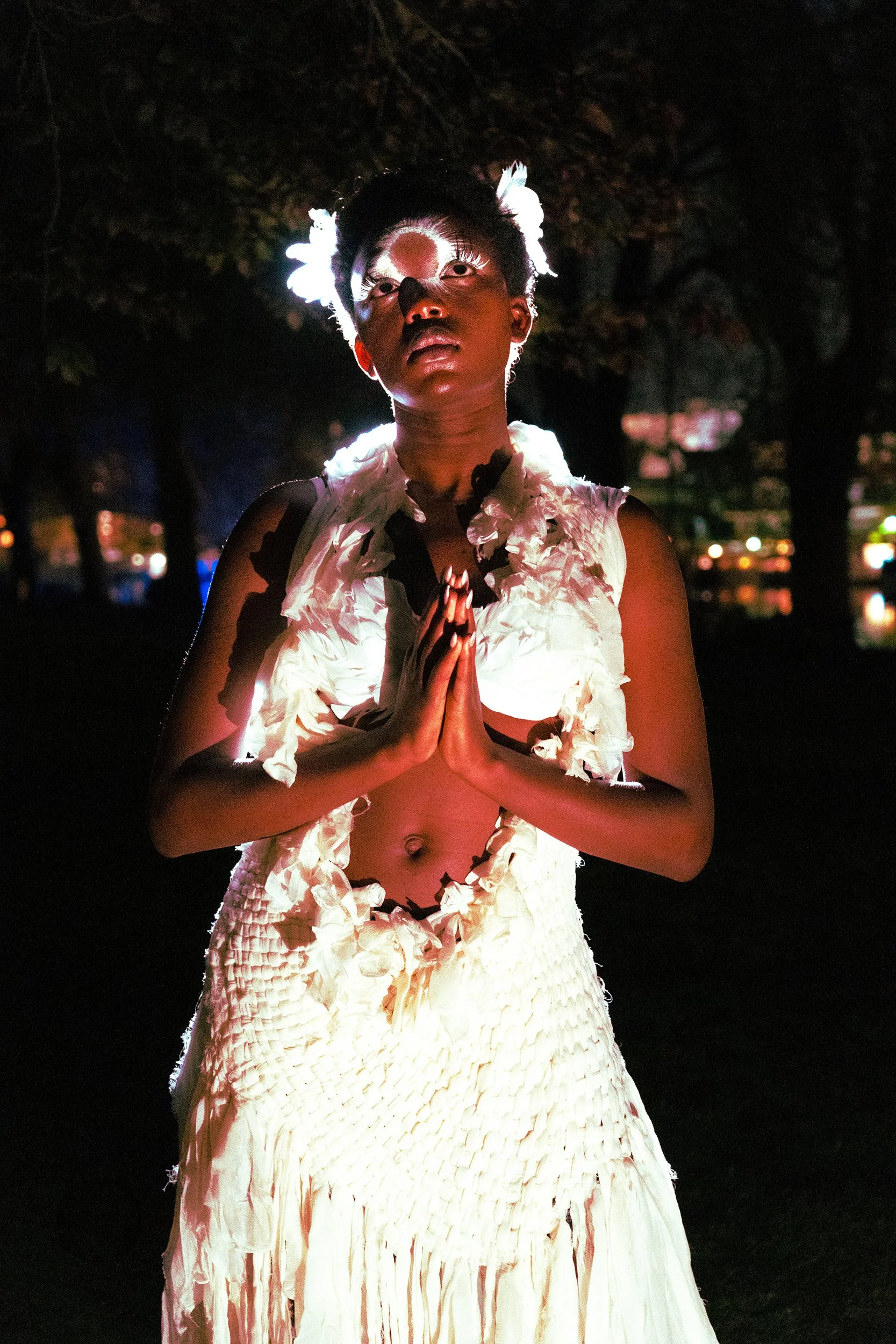 A person with makeup and white feathers on their head and chest, standing outdoors at night with hands pressed together in a prayer-like gesture, illuminated from below.