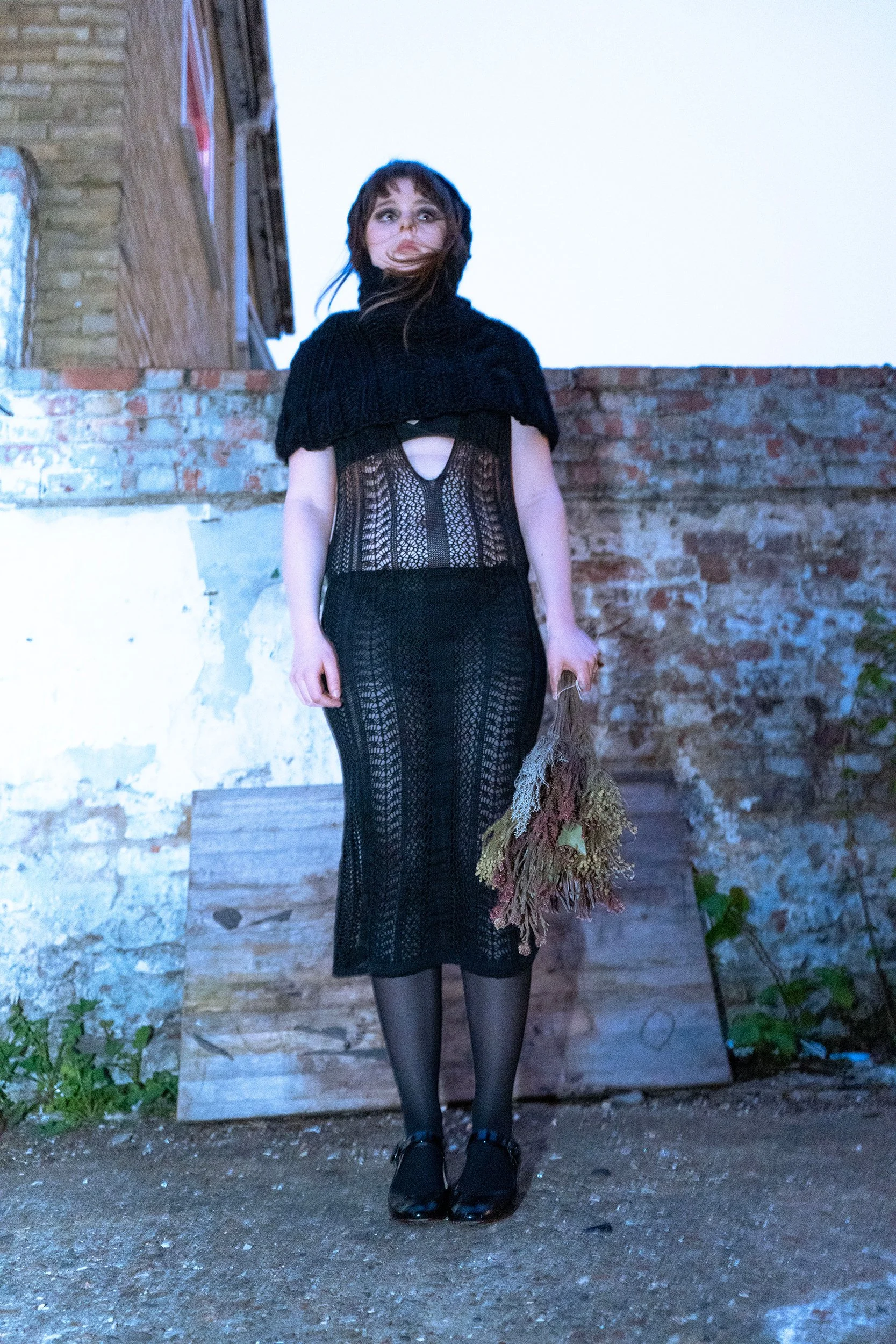 A woman with short brown hair stands outdoors against a brick wall, wearing a black crochet dress, stockings, and shoes, with a black shawl, holding a bouquet of dried flowers.