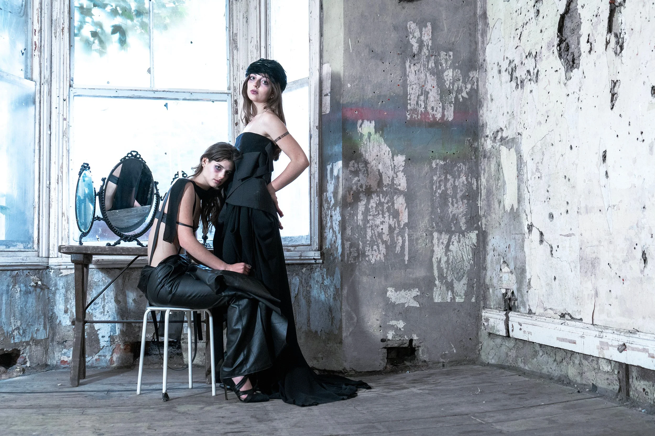 Two young women with dark hair and gothic fashion pose in an abandoned, grungy room with peeling paint and bare wooden floor. One woman stands with her hands on her hips, wearing a black off-shoulder dress and a black hat, looking to the side. The other woman sits at a small table with a vintage mirror, wearing black leather pants and a black top, resting her head on the table and looking directly at the camera.