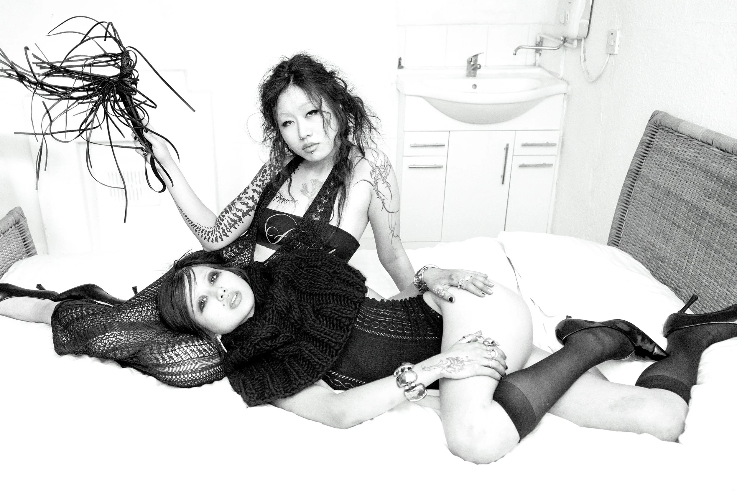 Two women posing on a bed in a minimalistic room. One woman is lying down wearing a black, see-through knitted top, and the other woman is sitting behind her with disheveled hair and tattoos. The room has a sink, faucet, and some shoes on the floor.