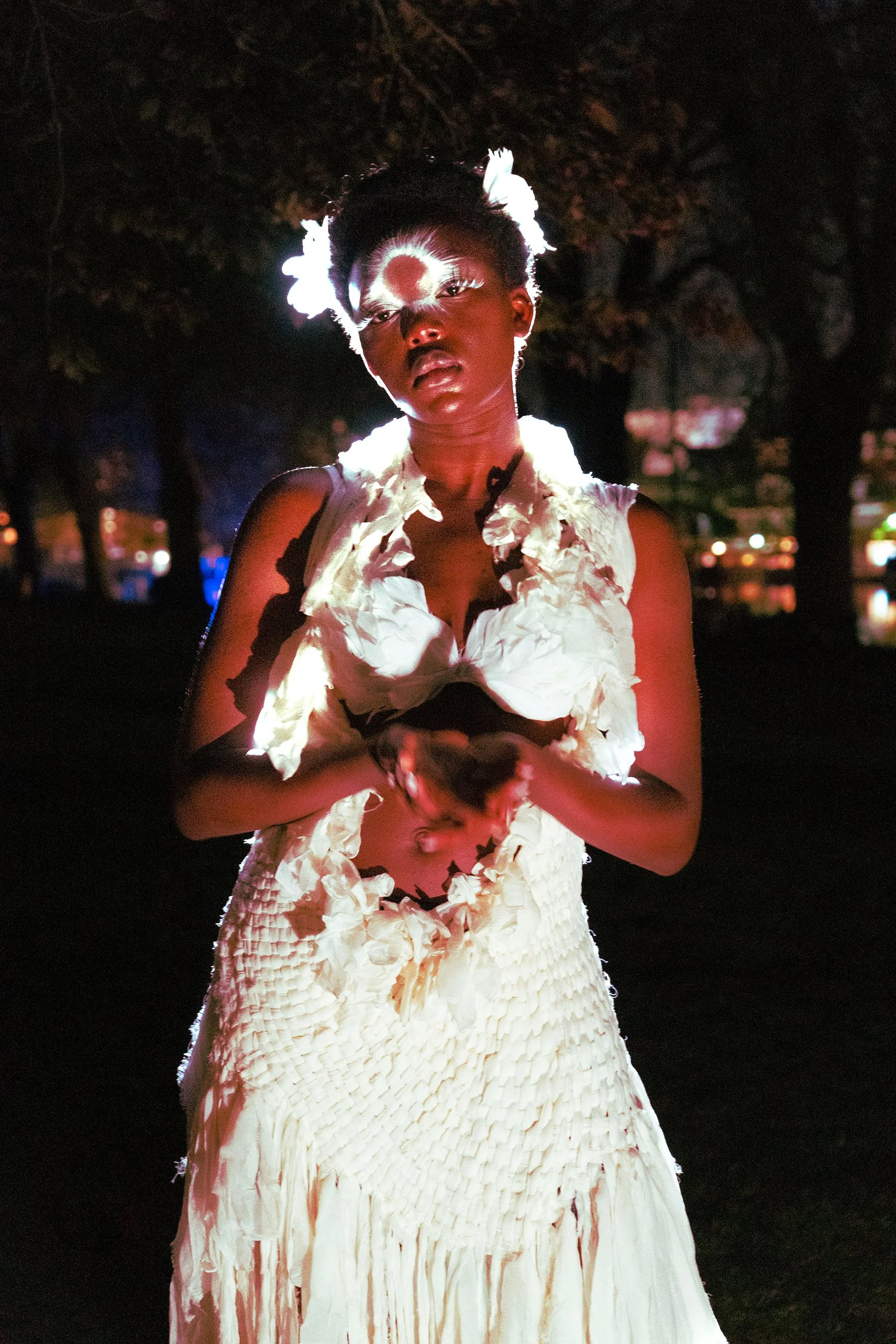 Night scene with a woman wearing a white, textured dress and white feathered accessories in her hair, standing outdoors with dark trees and blurred city lights in the background.