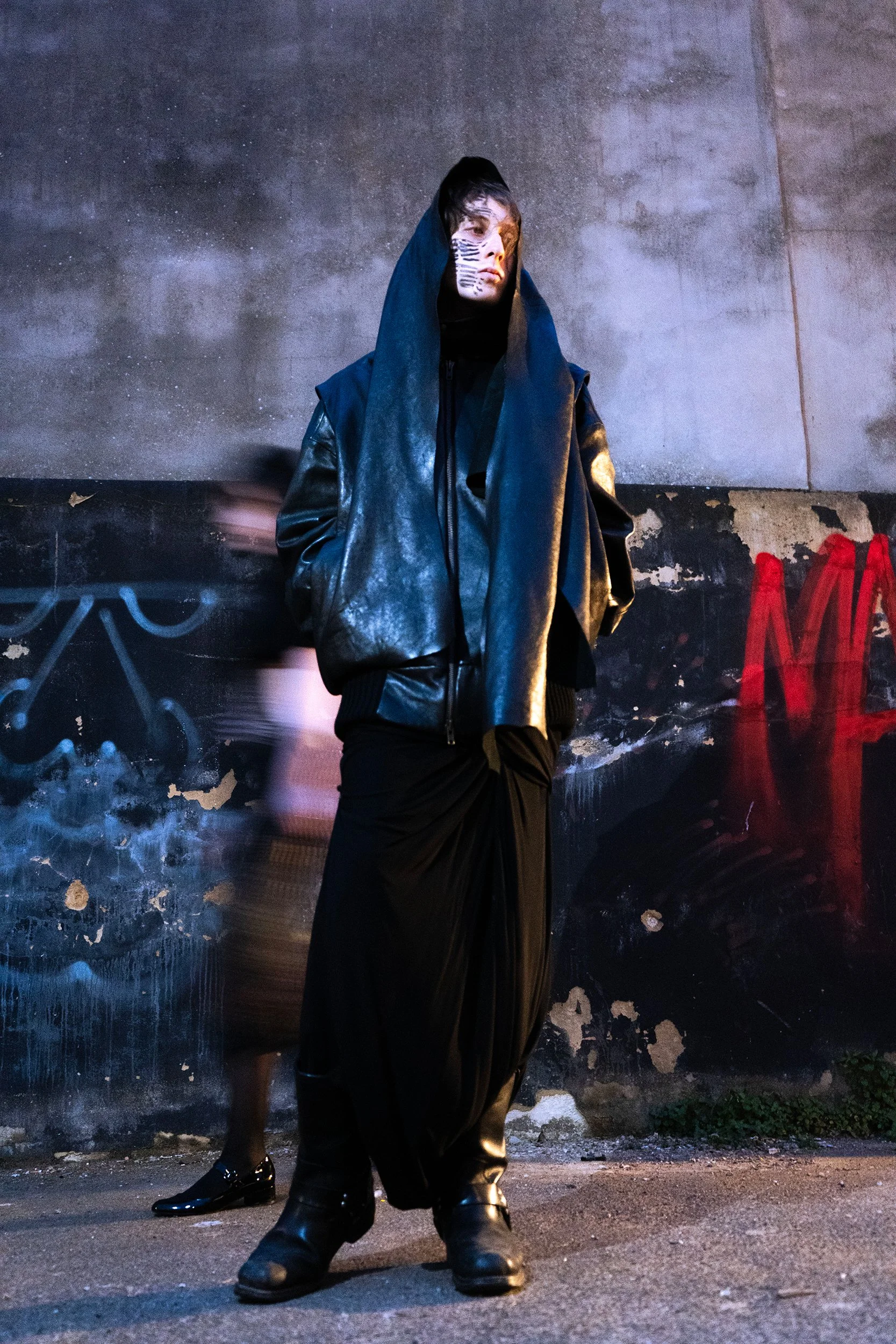 A person dressed in a long black coat and boots with a hood stands in front of a graffiti-covered wall. The person's face features zebra-like black and white painted stripes, and they are looking towards the camera. The background is urban, with a worn wall and some red graffiti on the right side. A blurred figure in black shoes is visible on the left side.