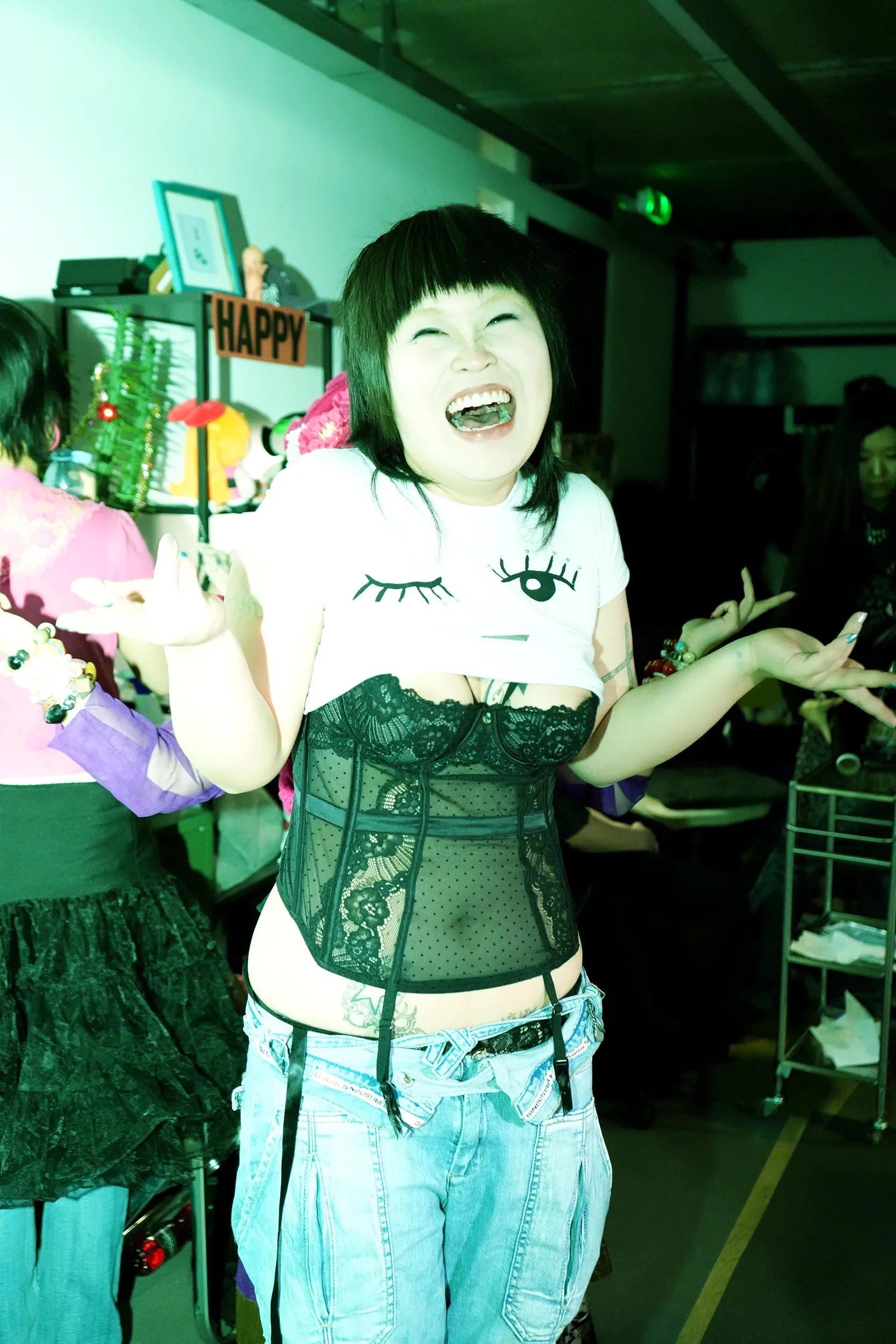 Person with short black hair wearing a black lace corset, white graphic t-shirt, and light blue jeans, smiling with mouth open in a party setting with decorations and other people in the background.