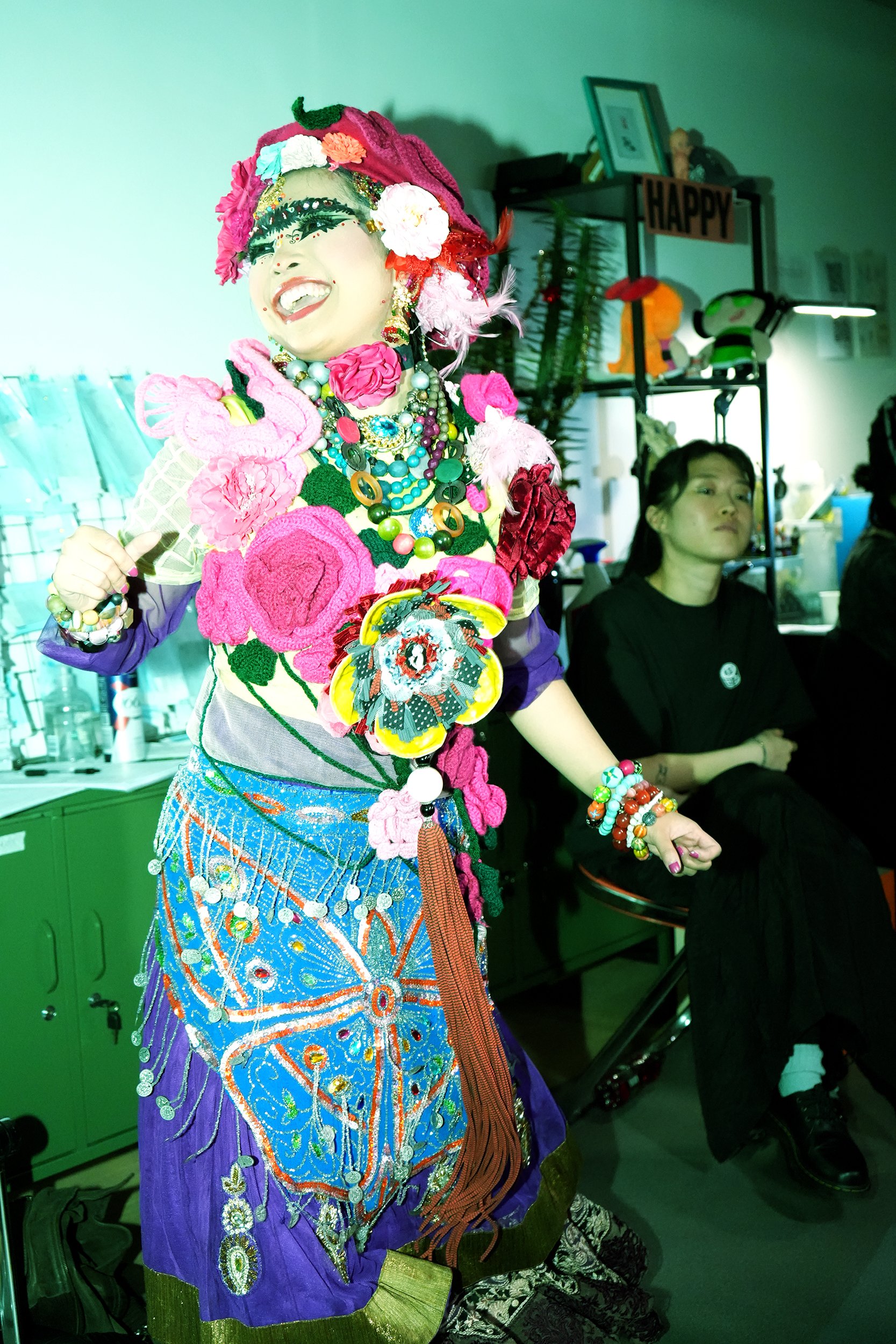 A person dressed in colorful, elaborate costume with floral patterns and jewelry, smiling and posing. In the background, another person sits, looking to the side, and a shelf with decorations and a sign that says 'HAPPY' is visible.