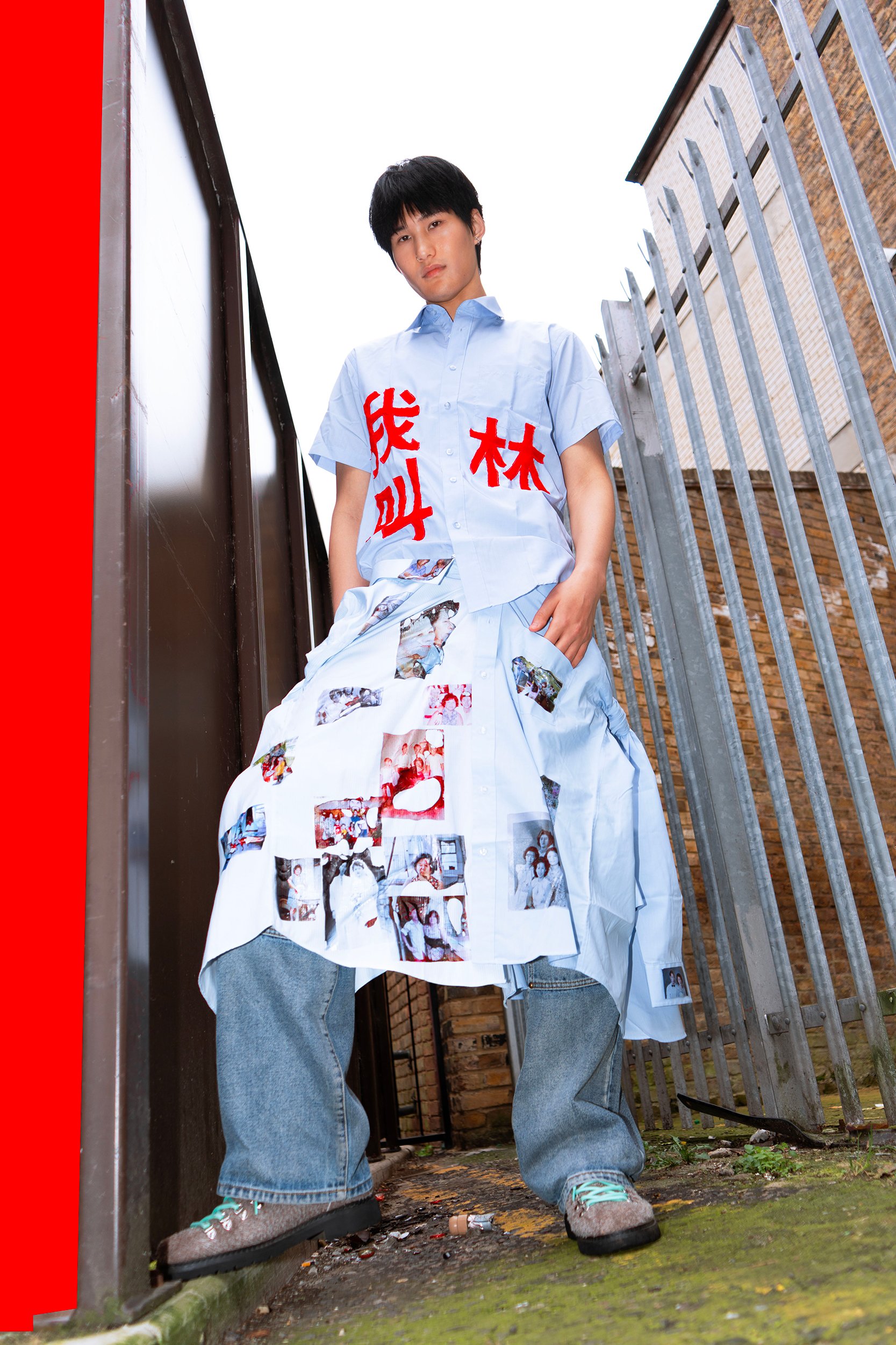 A young man standing outdoors between metal gates and brick walls, wearing a light blue shirt with red writing, a long white skirt with photos on it, and blue sneakers, looking at the camera.