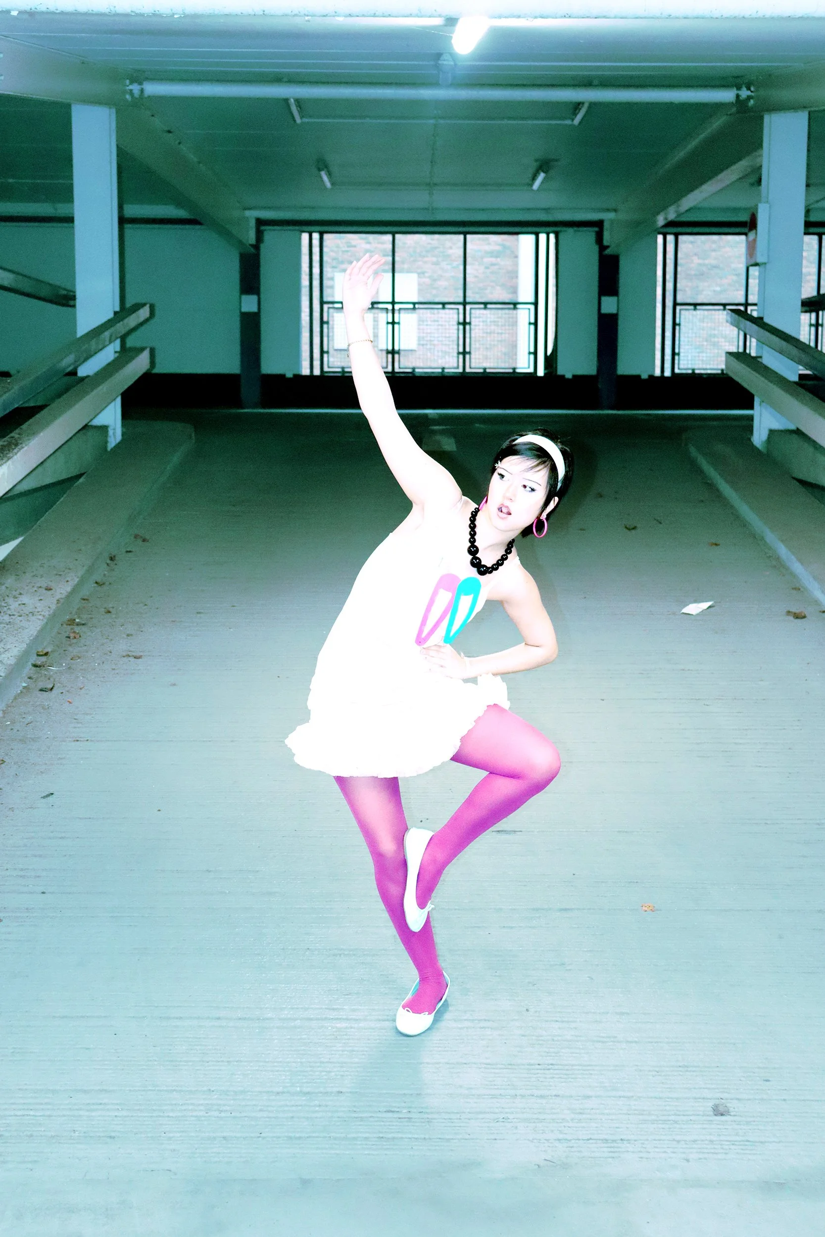 Person with black hair, wearing a white dress, pink tights, and white shoes, striking a pose in an indoor parking garage with large windows and railings.