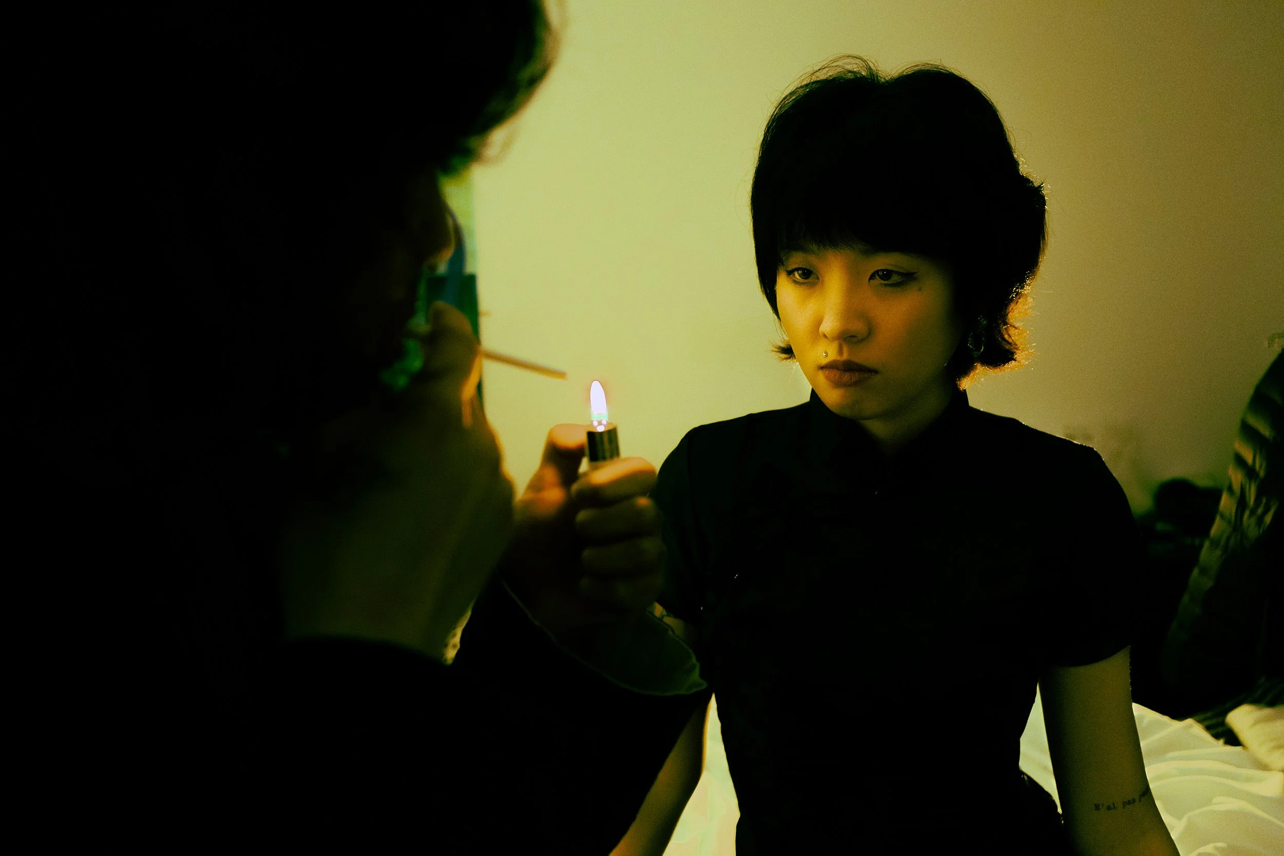 A young woman with short black hair looking pensively at a person off-camera while someone else holds a lit lighter close to her face.