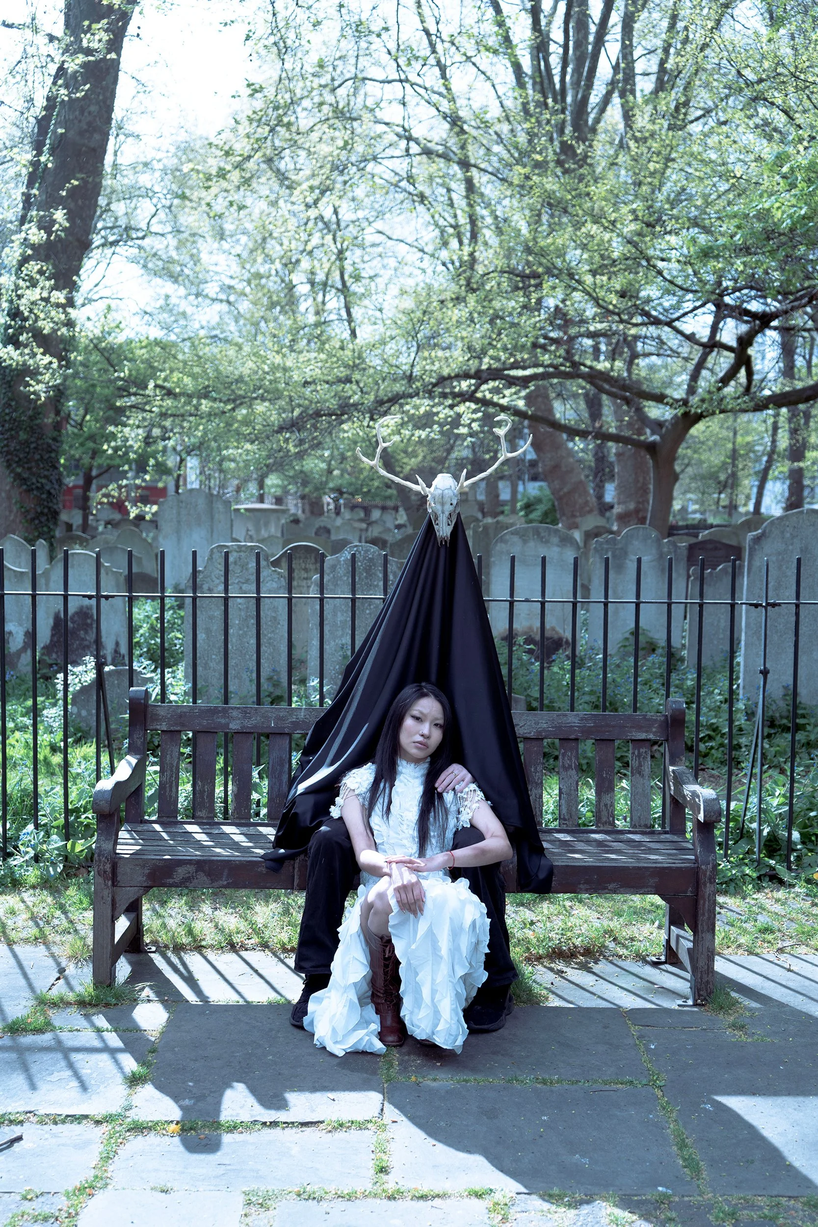 A young woman in a white dress and brown boots sits on a wooden park bench with her eyes closed and head tilted back. Behind her, a person dressed in black with a black cape, wearing a deer skull with antlers on their head, is seated, partially hidden by the cape. The background features a green, leafy park with trees, tombstones, and a metal fence.