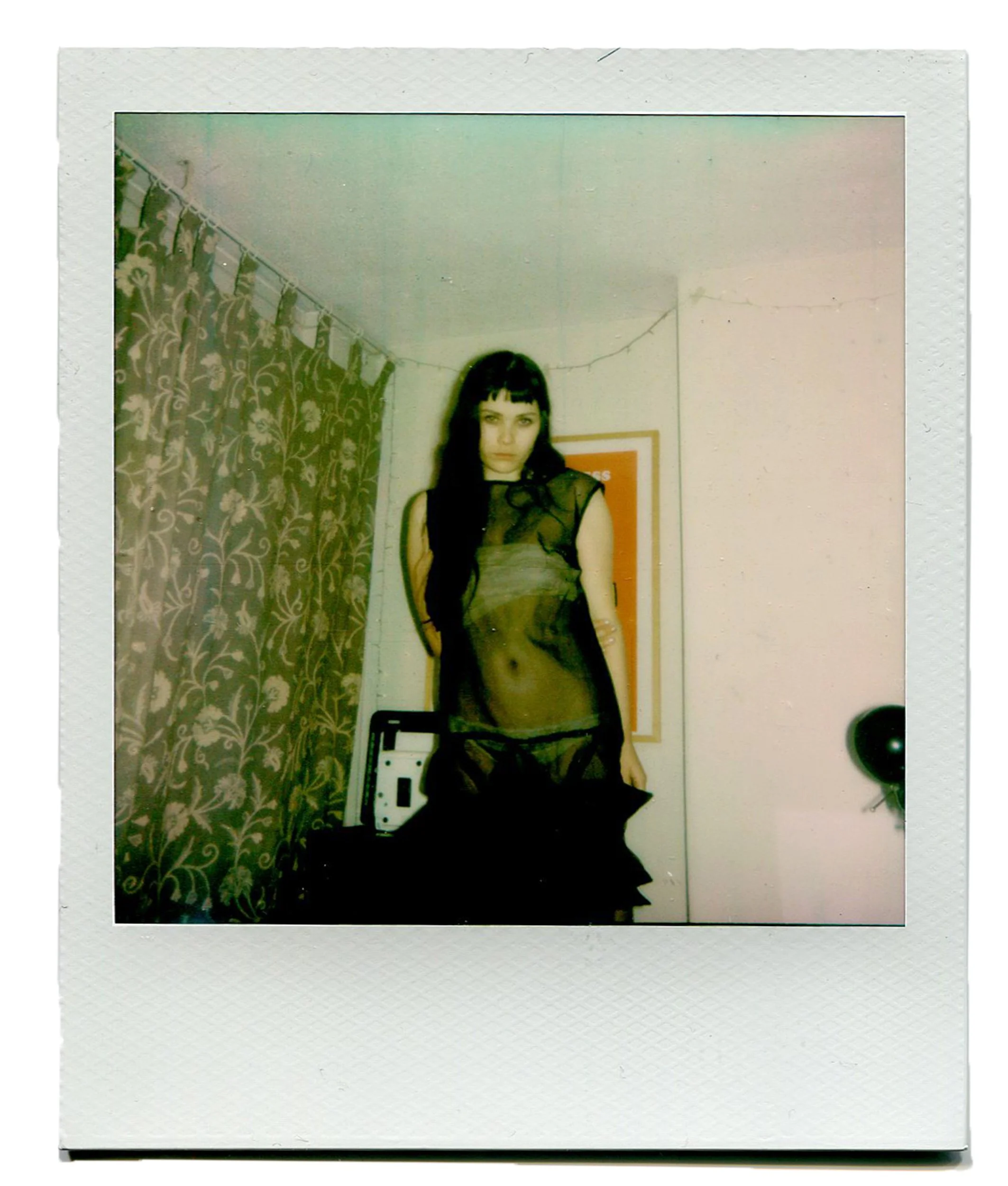 A young woman standing in front of a mirror, wearing a sheer black top and black underwear, with dark hair and bangs, in a room with patterned curtains and a string of lights on the ceiling.