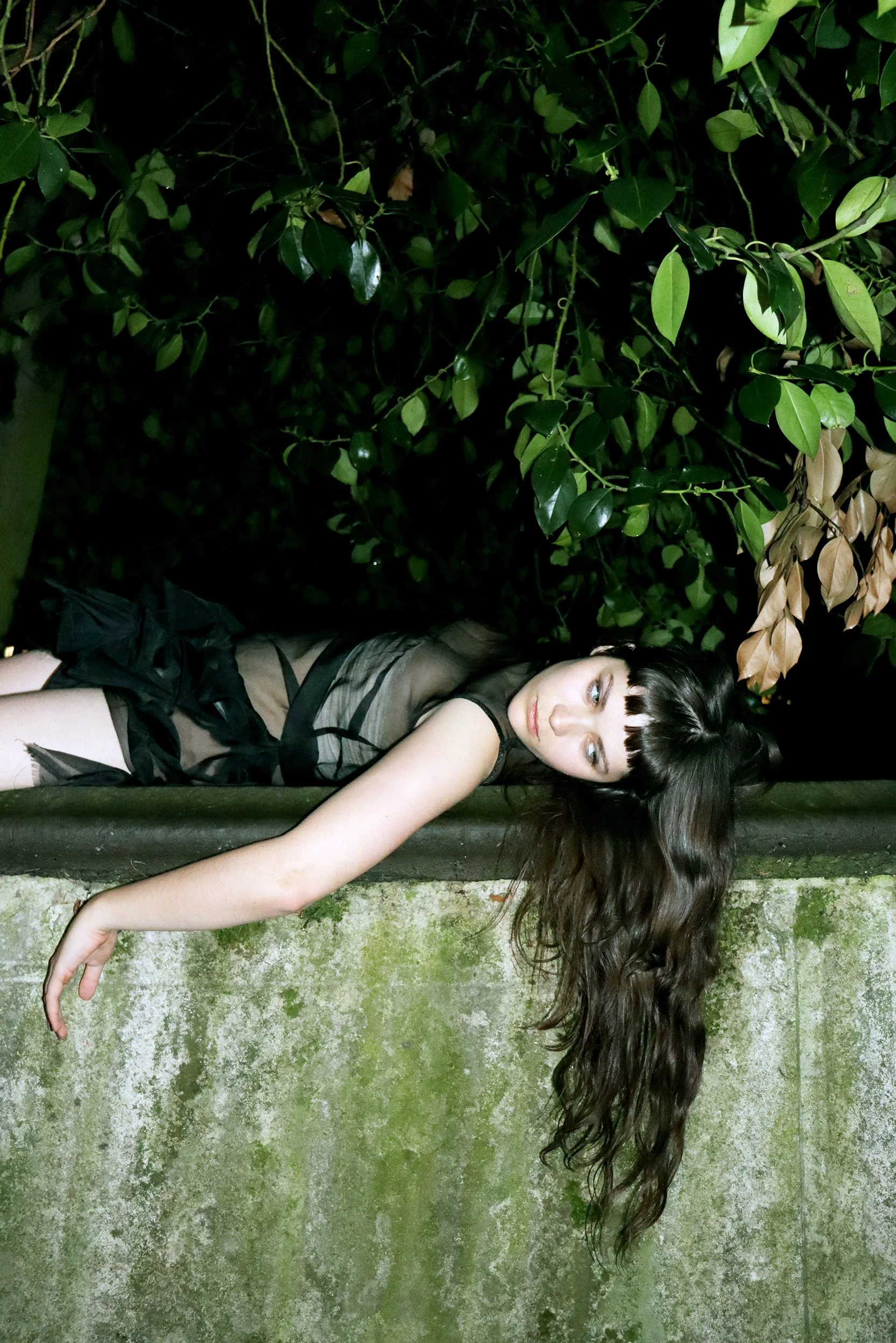 A woman with long dark hair, wearing a black sheer dress, laying upside down on a moss-covered stone ledge in front of dense green foliage at night.