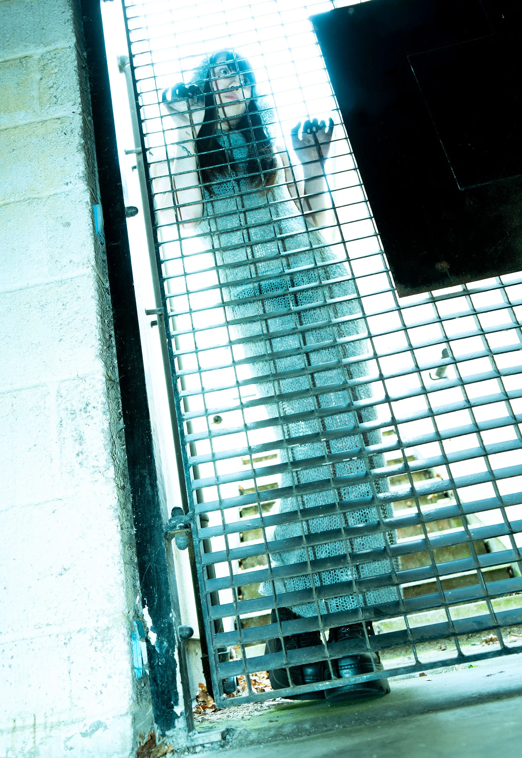 A woman with long dark hair and light skin appears behind a metal security gate, standing on a porch or driveway. She has a surprised or scared expression and is reaching out through the bars of the gate with one hand.