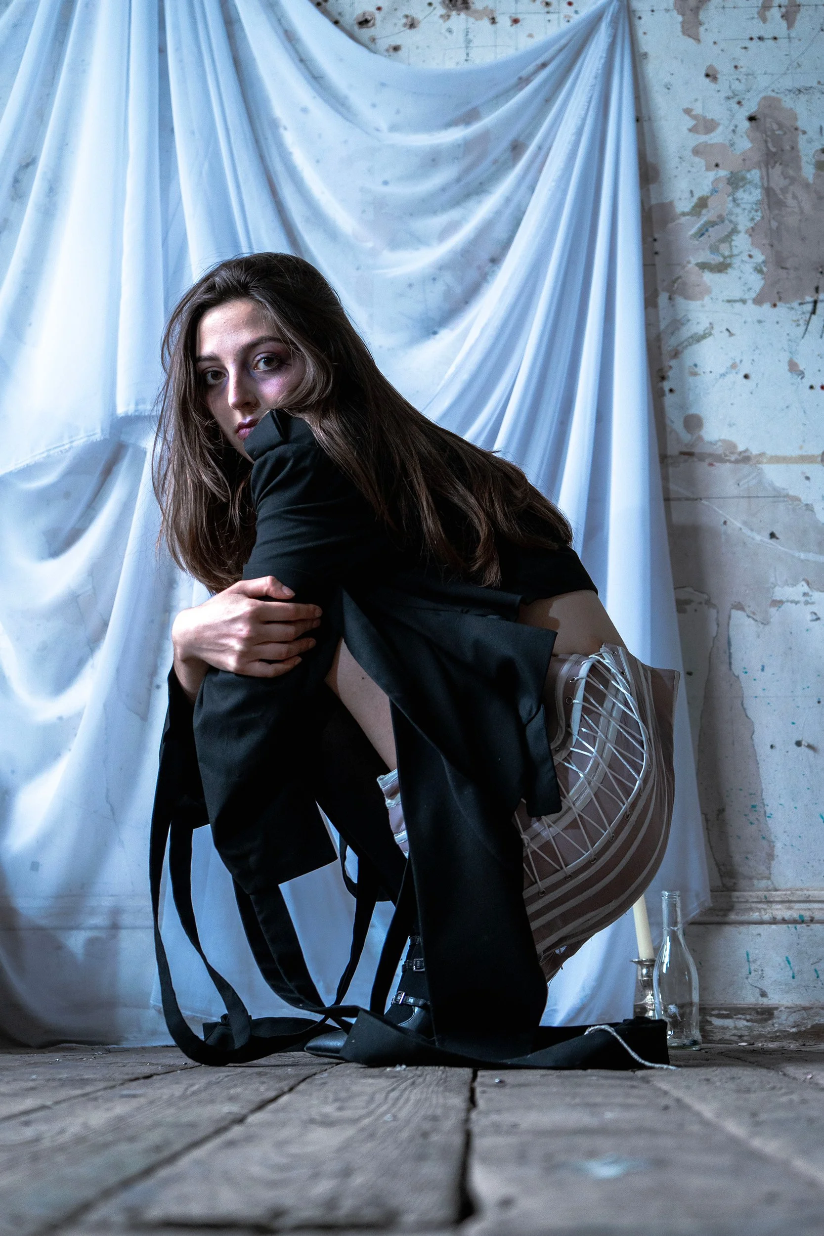Woman with long brown hair crouching on a wooden floor, holding her arm, in an industrial setting with a white draped fabric and a concrete wall in the background.