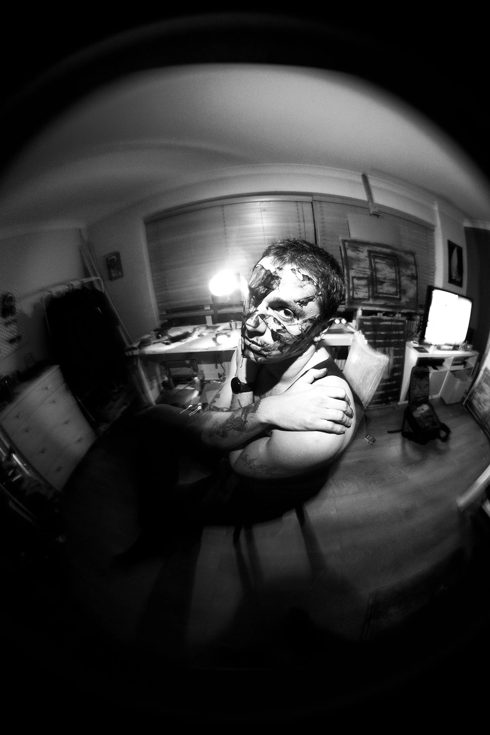A person with face paint resembling a spider or web design on their face takes a selfie in a room with computer desks, shelves, and a television, with a fisheye lens effect.