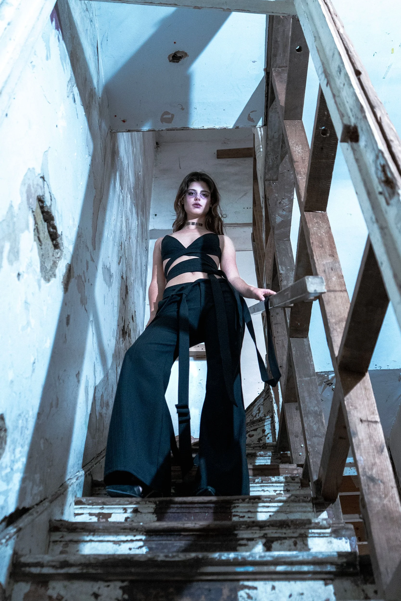 A woman with long wavy hair wearing a black cutout top and black pants standing on a worn wooden staircase in an abandoned, peeling-wabeled building.