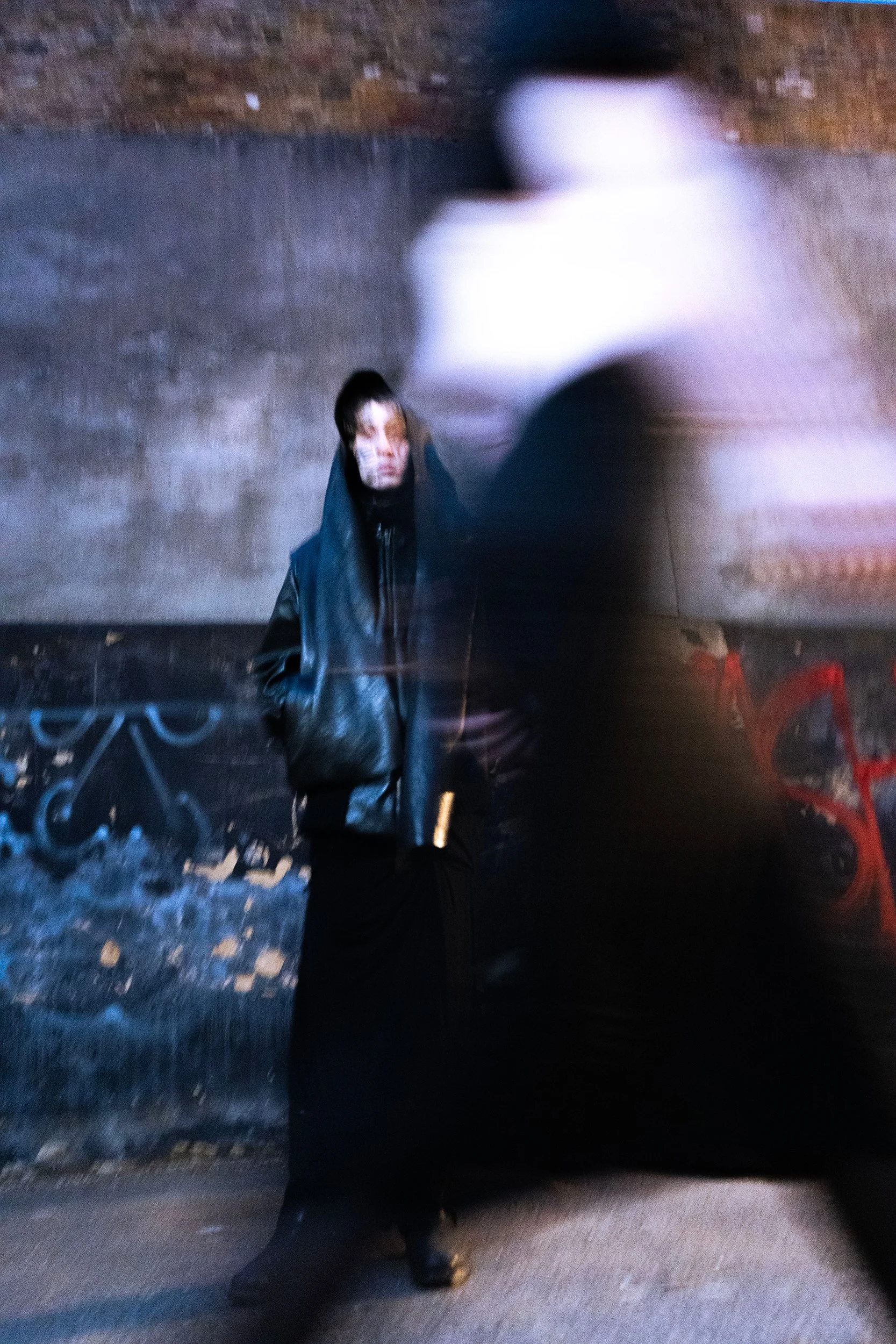 Blurred photo of a person in a dark jacket standing outdoors at night, with blurred figure of another person passing by in front of them, against a brick wall with graffiti.