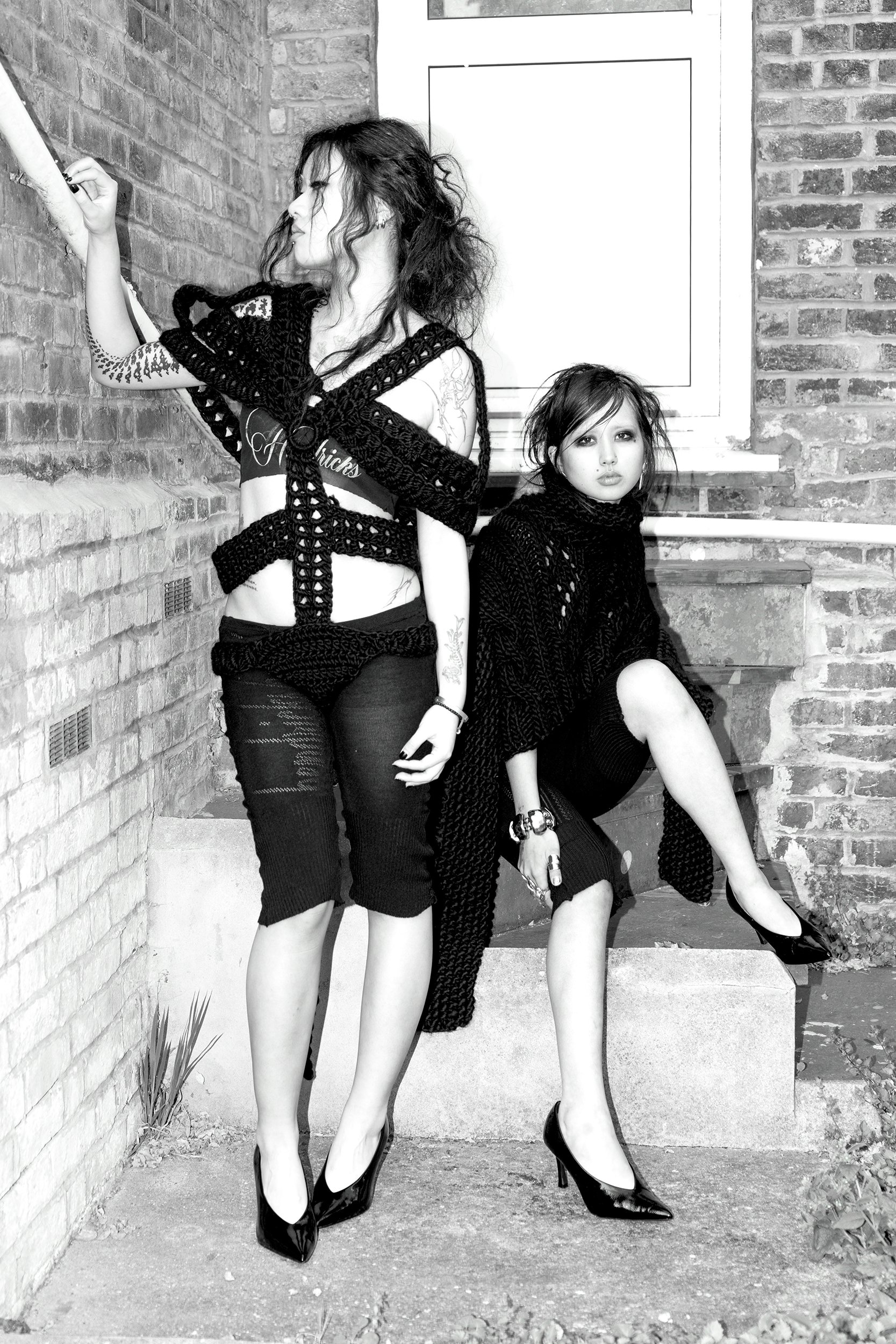 Two women with dark, wavy hair and pale skin, dressed in black, posing against a brick wall and a step outdoors. One woman is standing, the other is seated, both wearing high heels. The image is in black and white.
