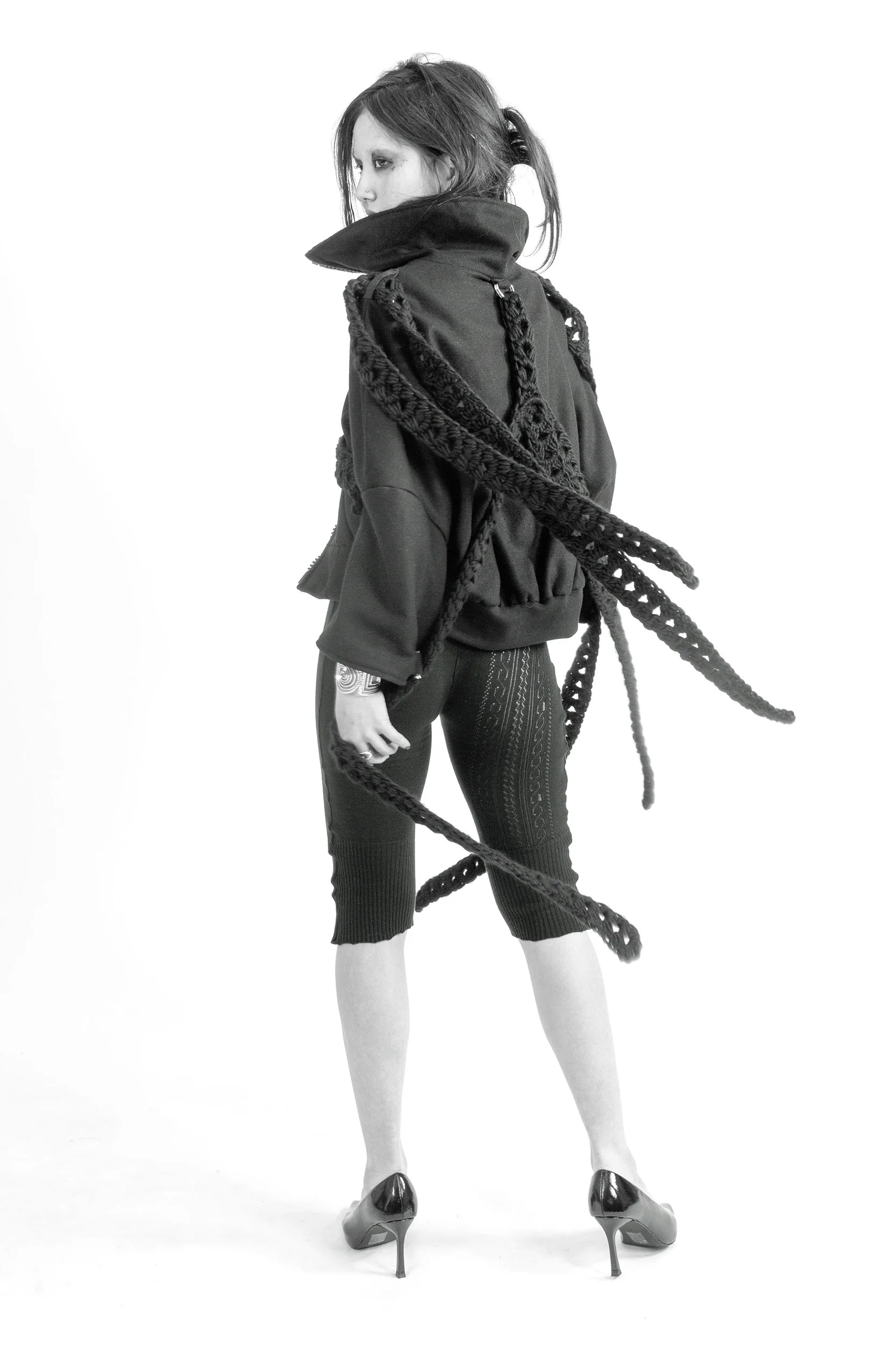 Black and white photo of a woman with shoulder-length hair wearing a large jacket, knit scarf, patterned leggings, and high heels. She is standing against a plain white background, facing slightly to the left with her head turned to the side.