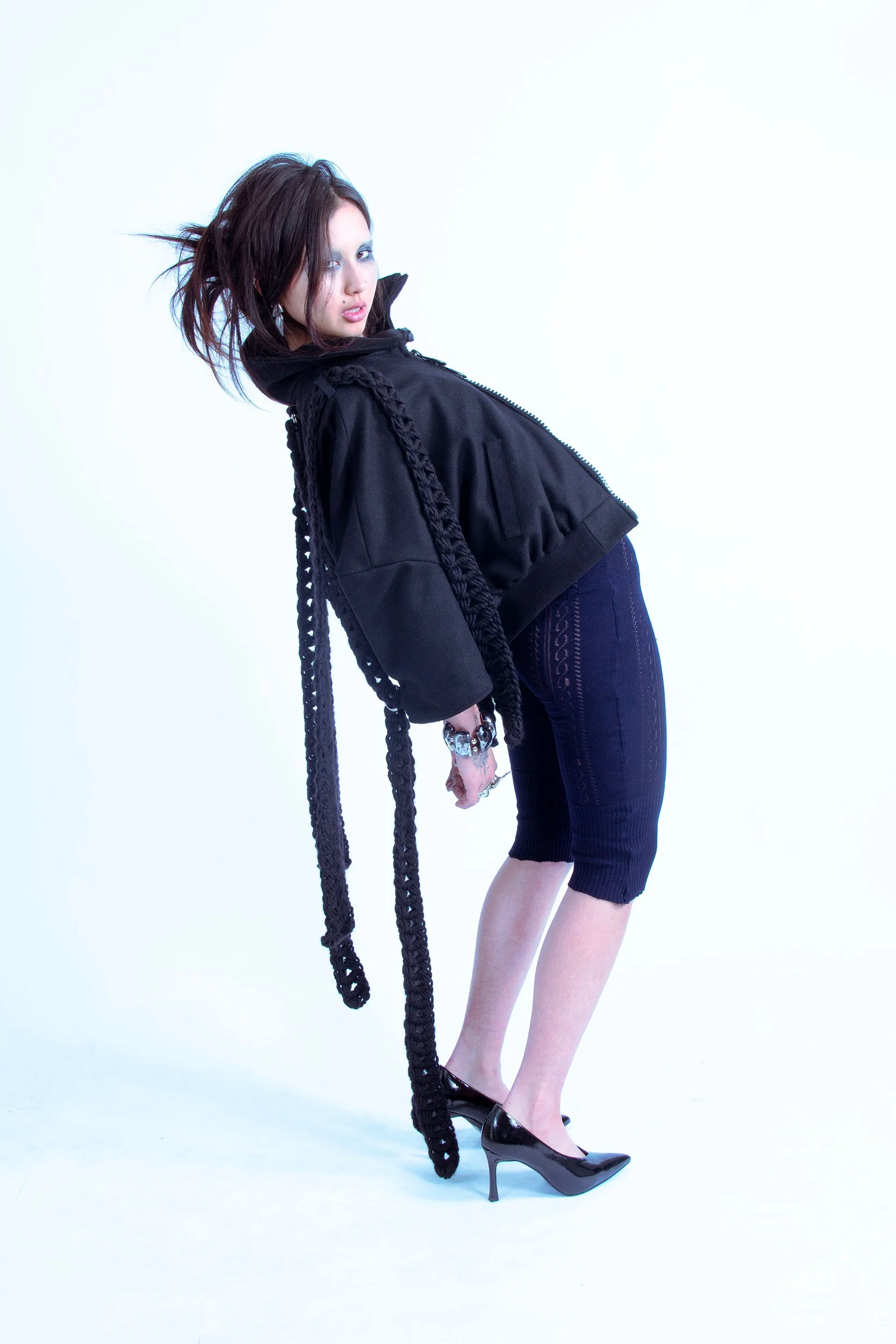 A woman with short, messy brown hair, wearing a black jacket, navy blue dress, and black heels, is posing against a white background with her body bent forward and her head turned to look at the camera.