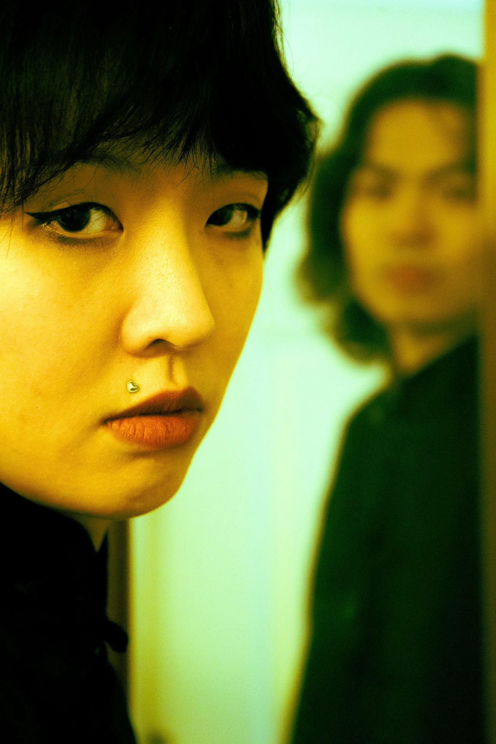 Close-up of a young woman's face with short black hair, winged eyeliner, and a septum piercing, with another woman blurred in the background.
