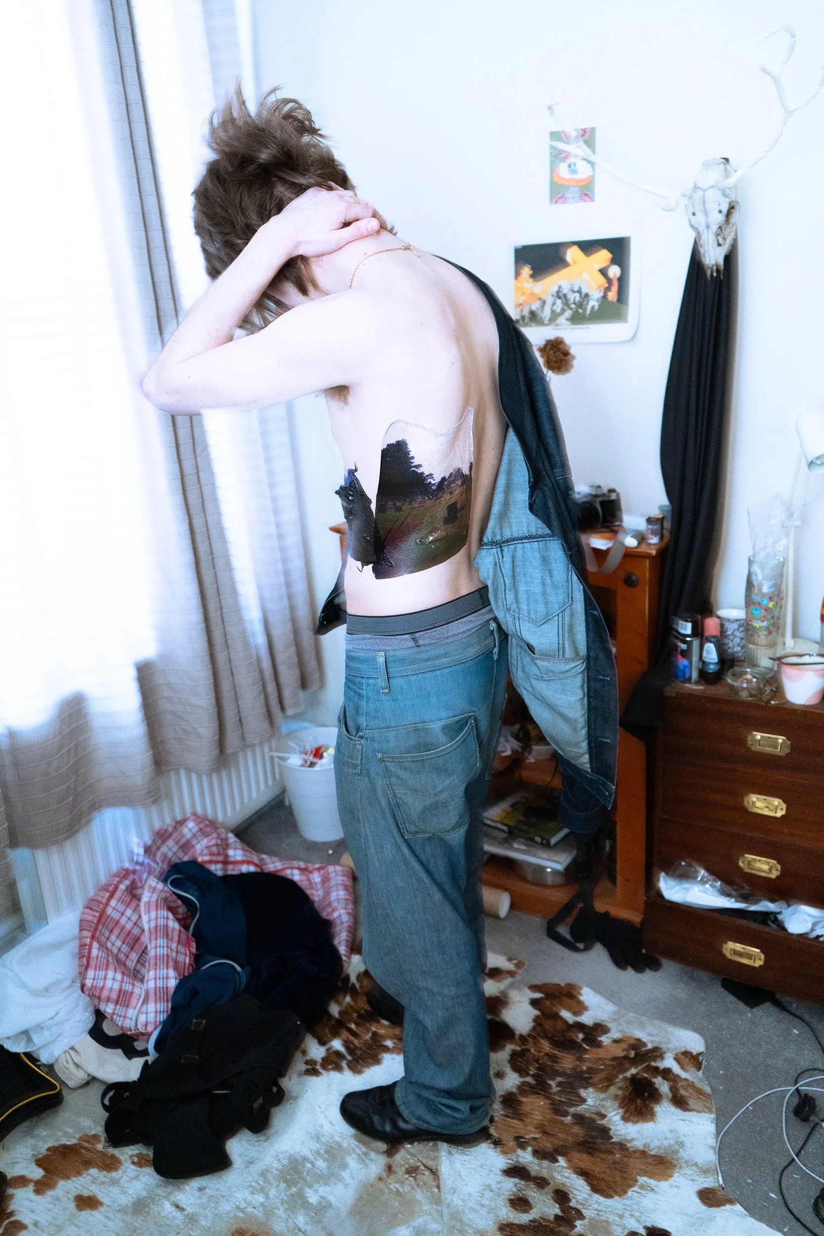 A person with short brown hair, wearing blue jeans, is standing in a cluttered room. They are topless, with a painted landscape on their side. The room has a cowhide rug on the floor, a dresser with various items, and art on the walls. There are clothes and backpacks on the floor, and a bucket near the window.