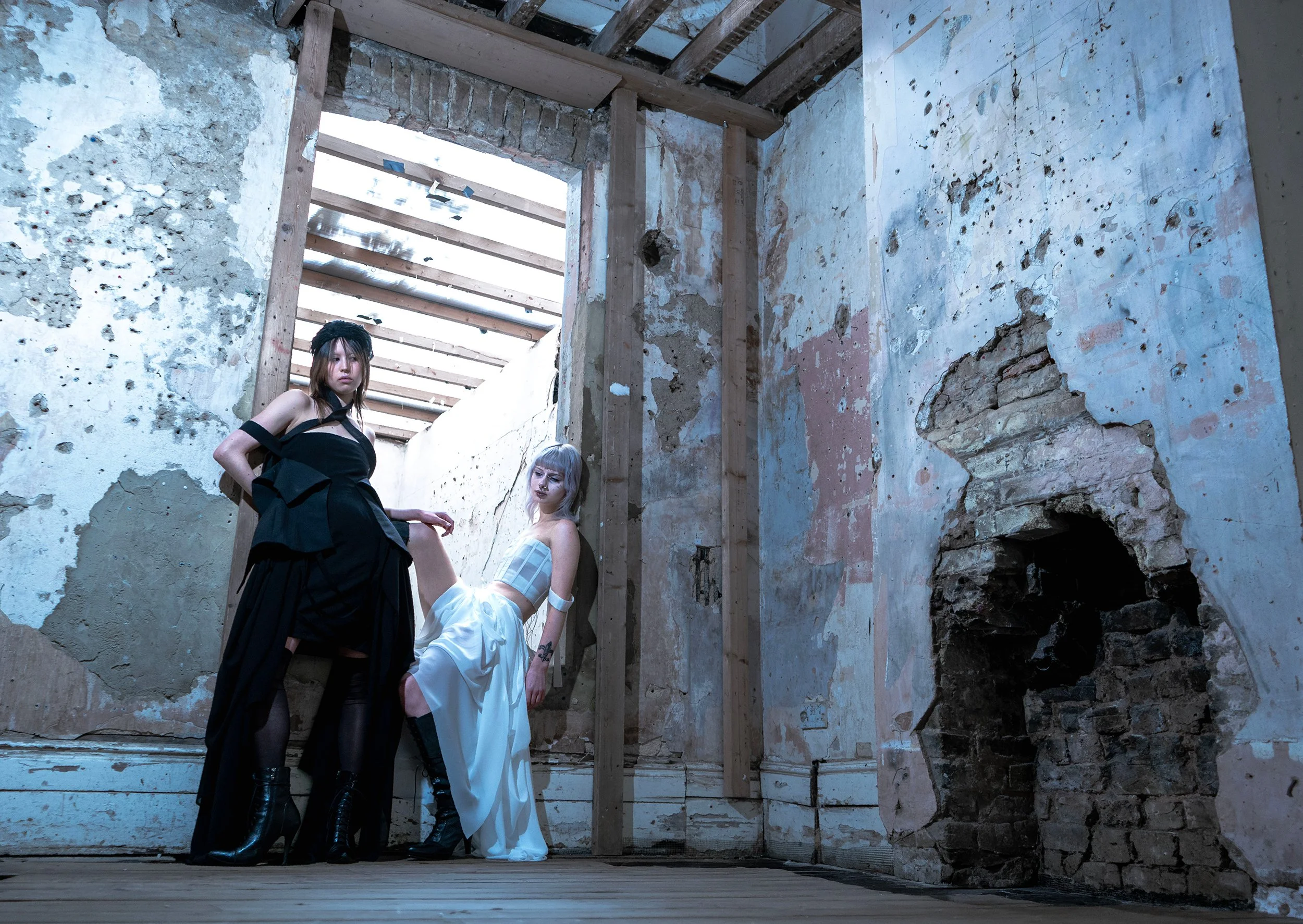 Two women in fashionable black and white outfits posing in a dilapidated building with exposed brick and wood beams.