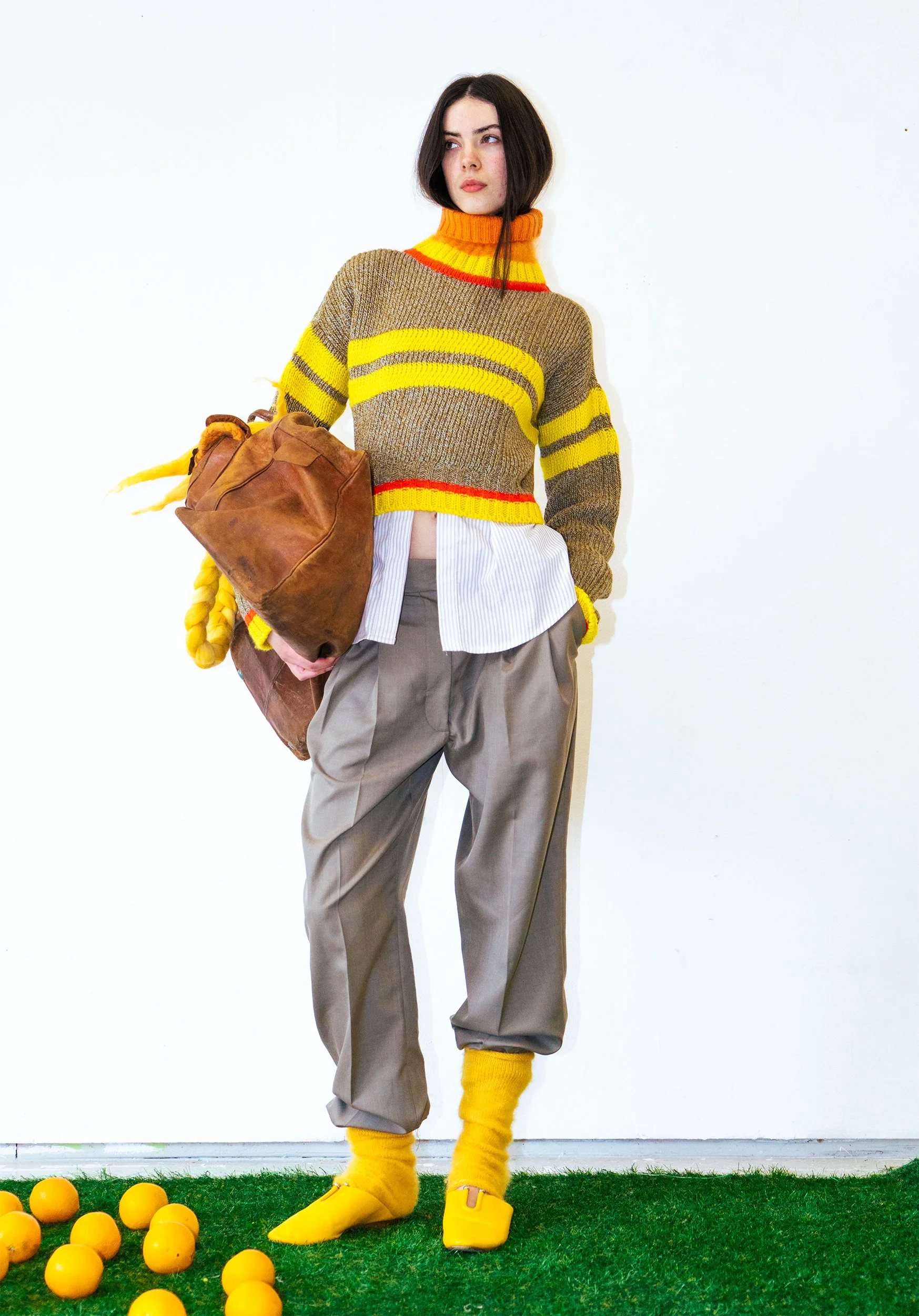Woman standing on artificial grass with yellow balls, dressed in a multicolored sweater, brown trousers, yellow socks, and yellow shoes, against a plain white wall.