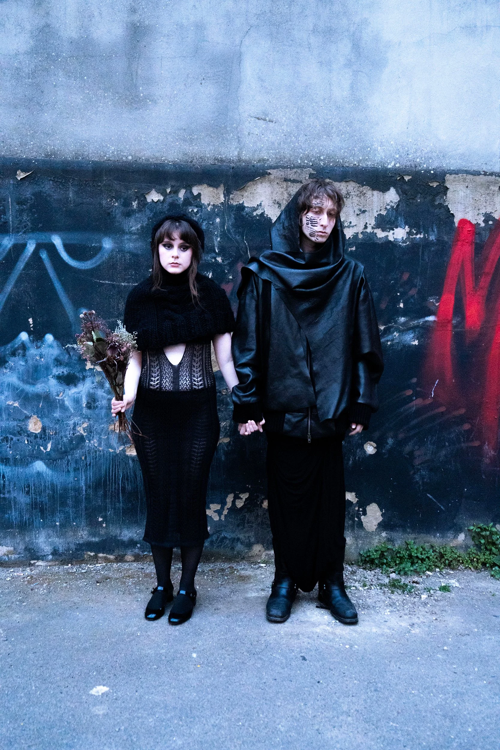 A woman with long brown hair wearing an all-black outfit holding a bouquet of flowers, standing next to a man with curly hair and face tattoos, dressed in a black hooded leather cloak and black pants, standing against a weathered graffiti-covered wall.