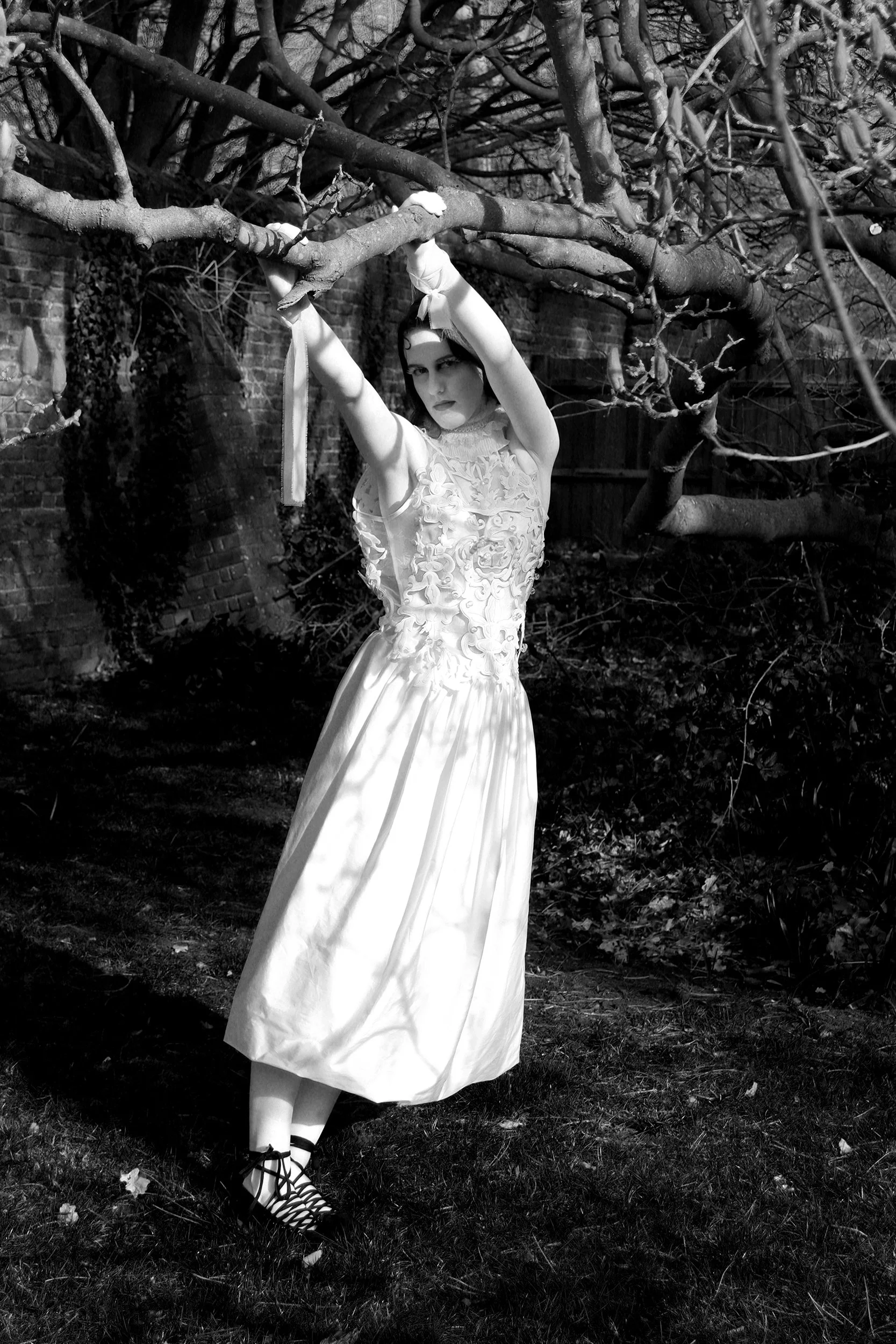 A woman in a long, elegant white dress and high heels stands outside, holding onto a large, leafless tree branch. The scene appears to be in a garden or backyard with a brick wall in the background. The photo is in black and white, giving it a dramatic, artistic feel.