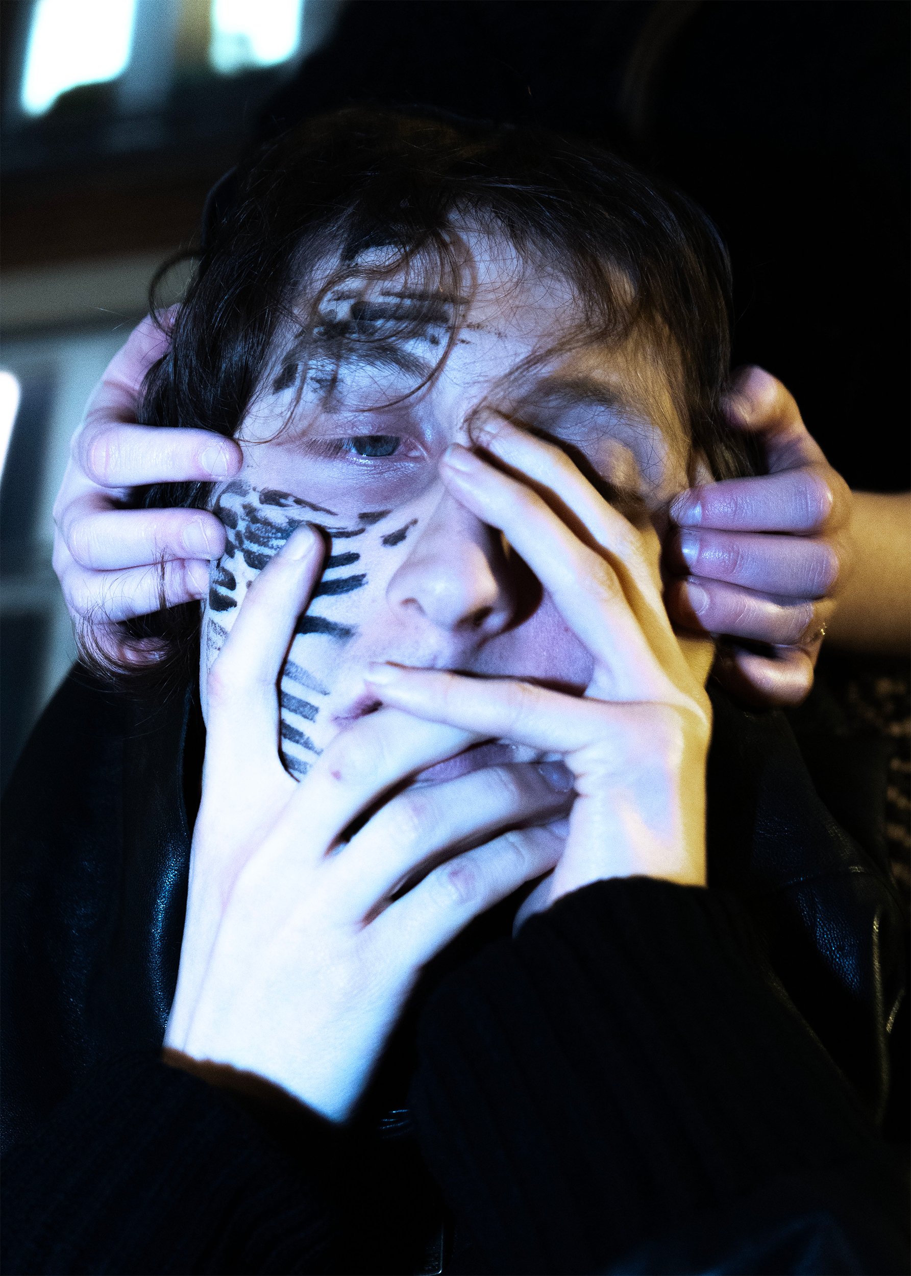 Close-up of a person with dark hair holding a tiger-striped cloth over part of their face and covering one eye, with hands near their face, in a dimly lit setting.