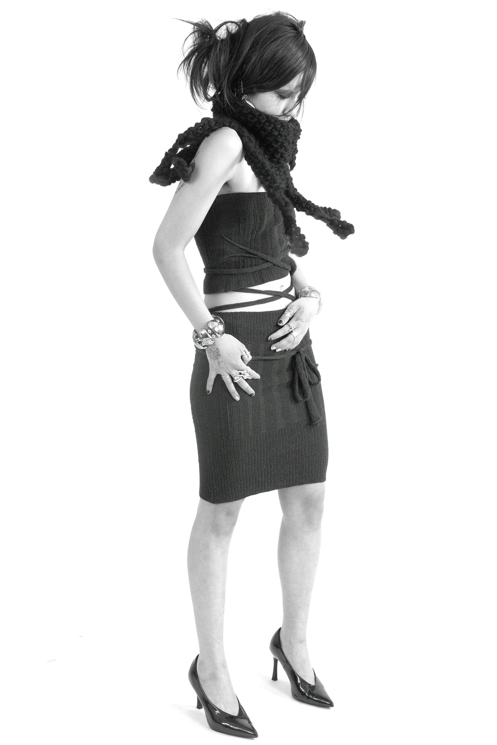 A woman dressed in a sleeveless black knit top and a fitted, ribbed black skirt with side slits, wearing black high heels, and accessorized with multiple bracelets and rings, standing in profile with her head bowed down and hands resting on her hips.