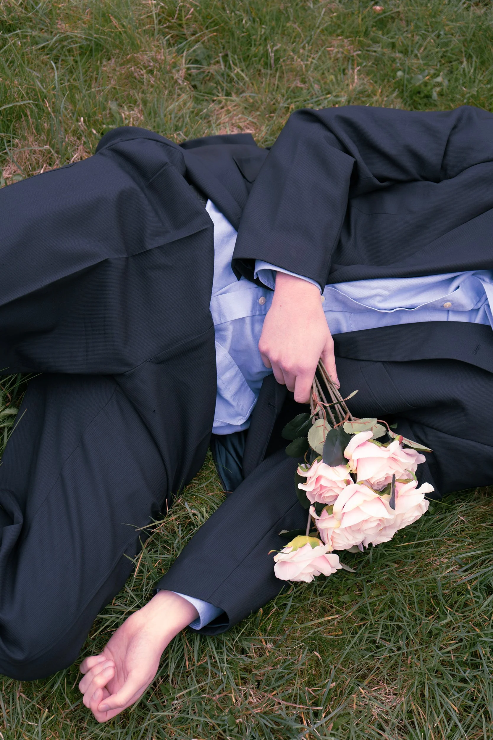 Person lying on grass in a black suit holding a bouquet of pink roses.