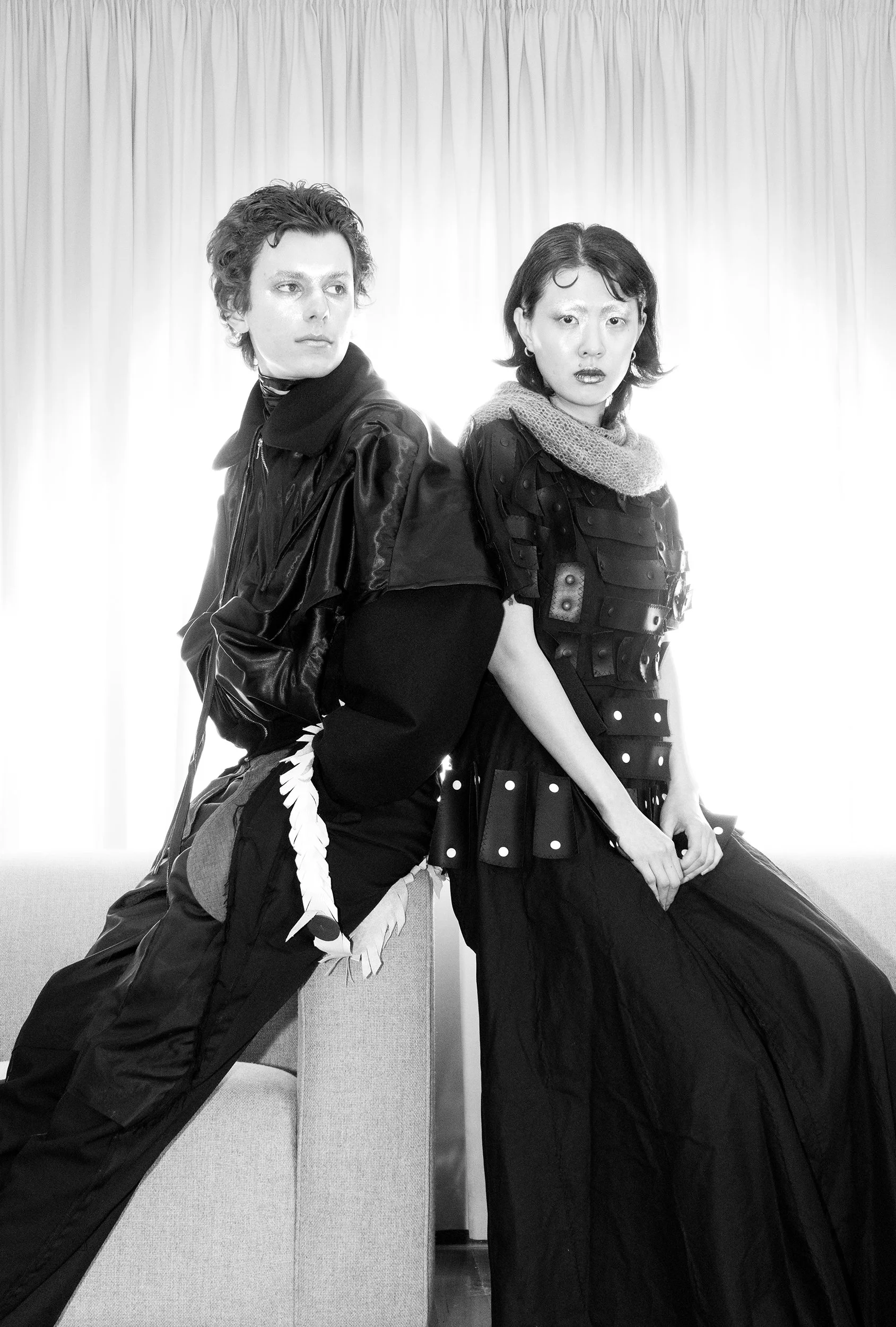Two models, one male and one female, posing in front of a light-colored curtain, dressed in dark, edgy clothing.