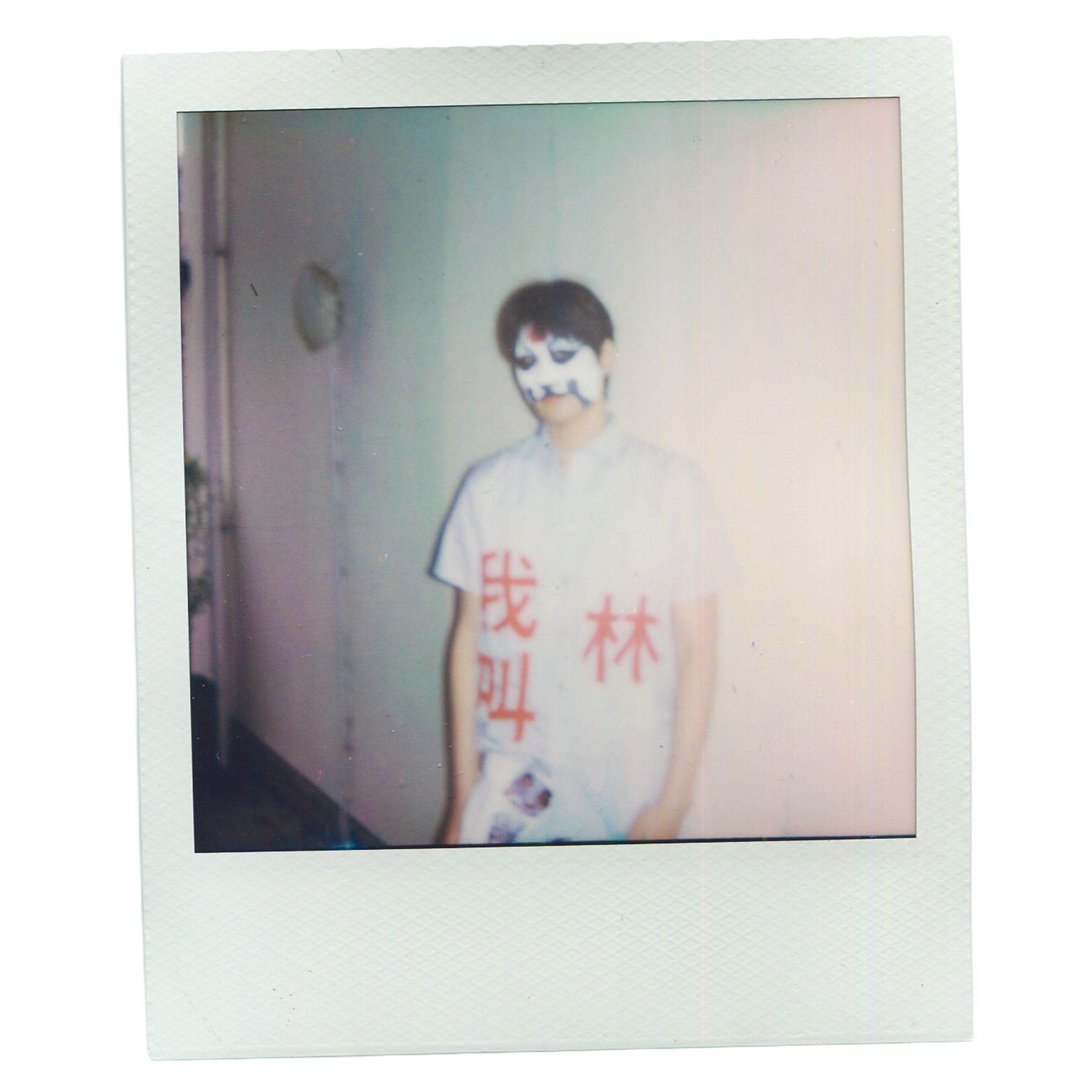 Polaroid picture of a person with short dark hair wearing a face mask, jewelry, and a beige shirt. The Polaroid has a white border, and the background appears to be an indoor setting.