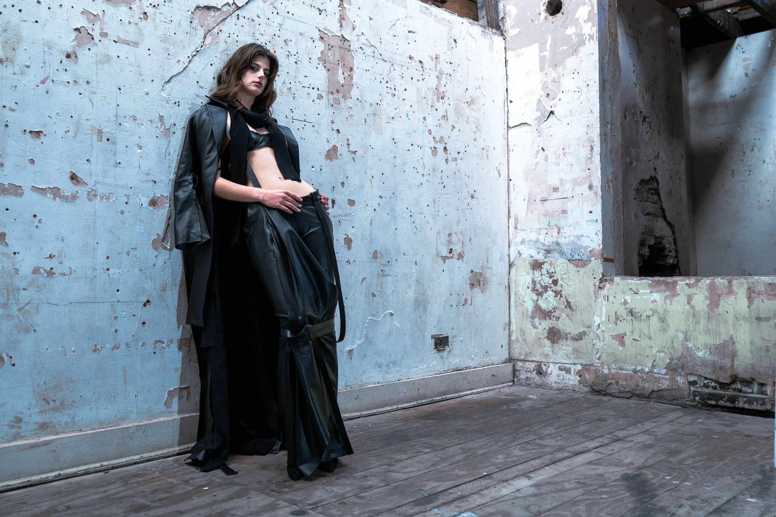 A young woman with shoulder-length brown hair leaning against a weathered, peeling painted wall in an abandoned or industrial space. She is dressed in a black leather jacket, black pants, and has a black scarf, with a serious expression on her face.