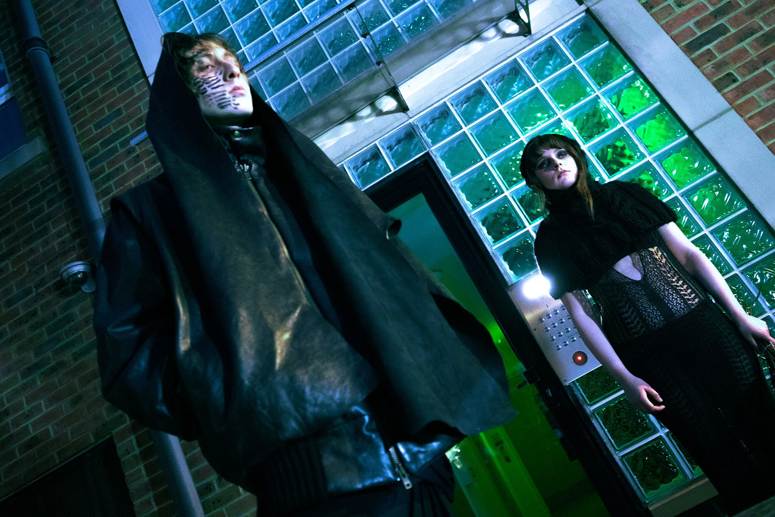 Two young women standing in front of a glass block wall with green lighting, one wearing a black hoodie and the other wearing a black lace dress.